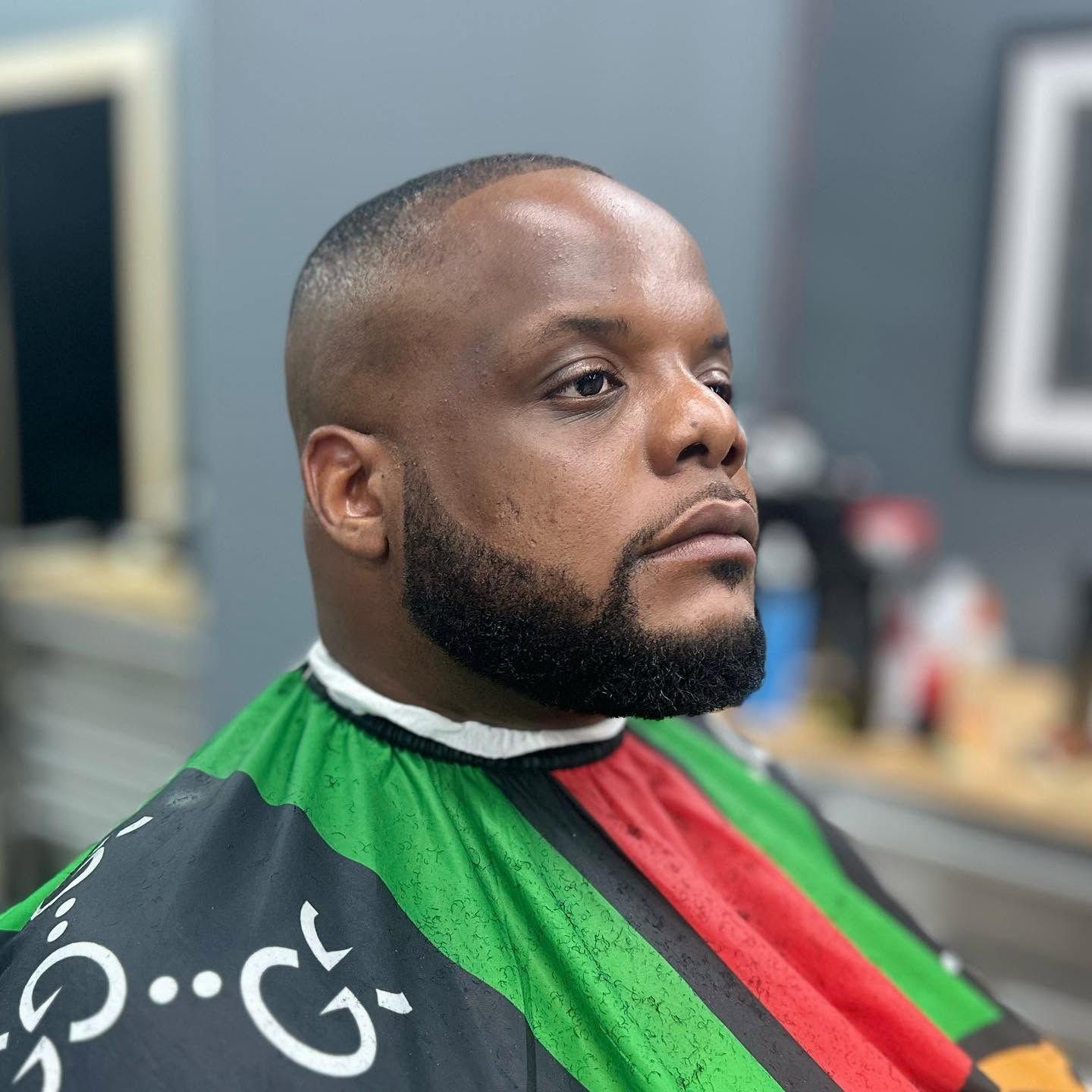 Man in a barber cape with green, red, and black stripes, seated in a barbershop, looking to the side.