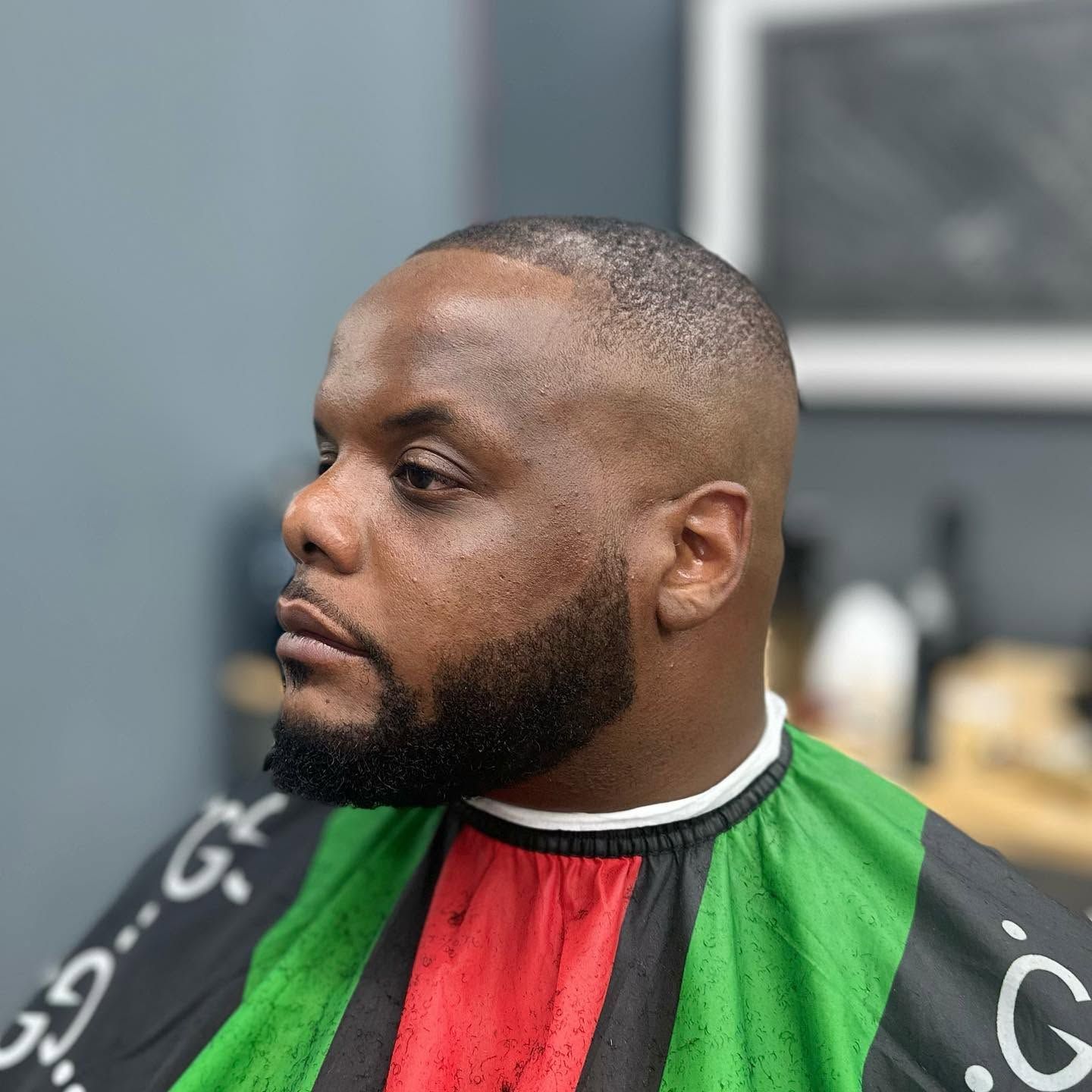 Man with a close-cropped beard wearing a red, green, and black barber cape, shown in profile.