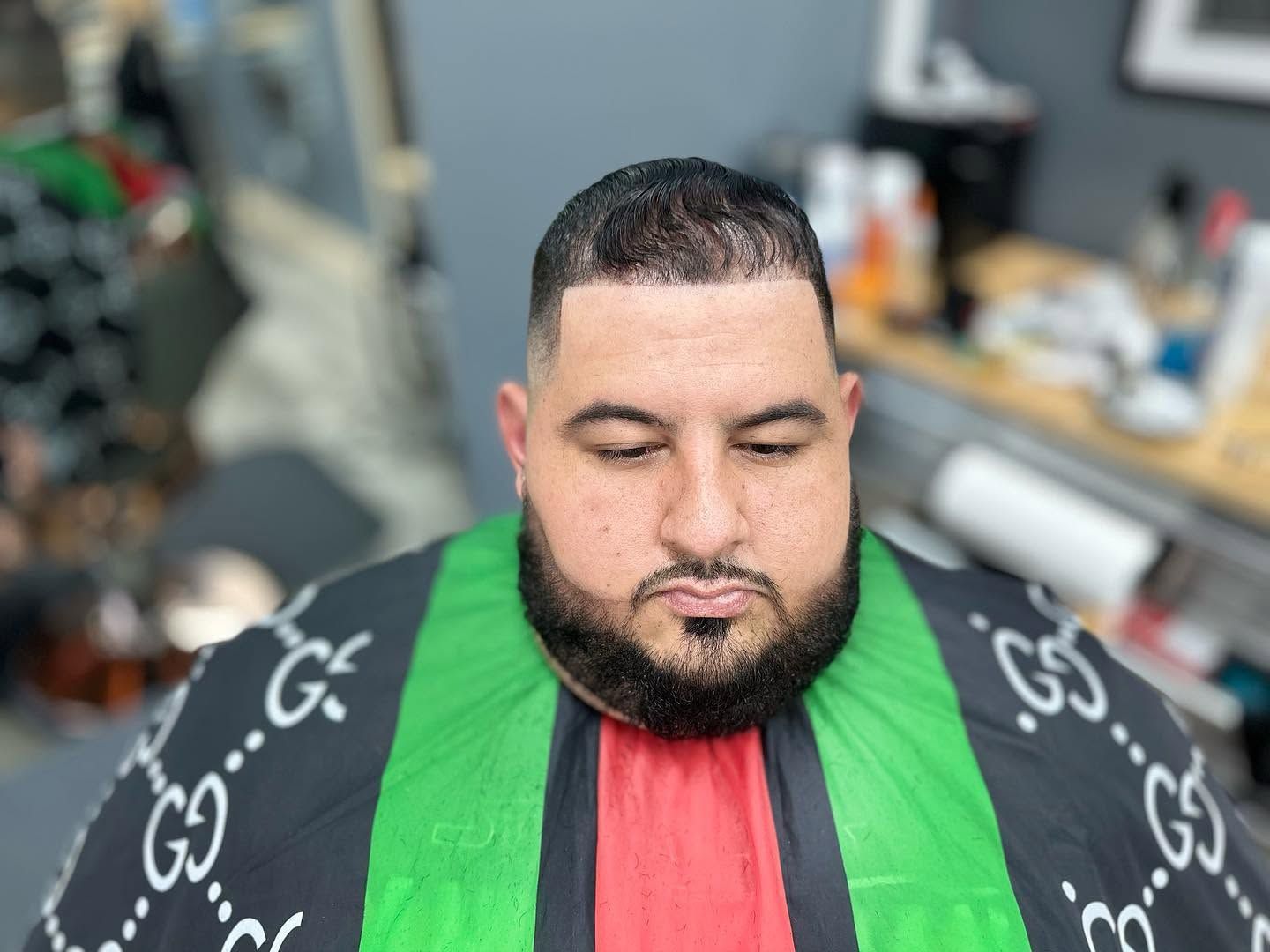 Man seated in a barber chair wearing a green, red, and black cape, with a fresh haircut and beard trim