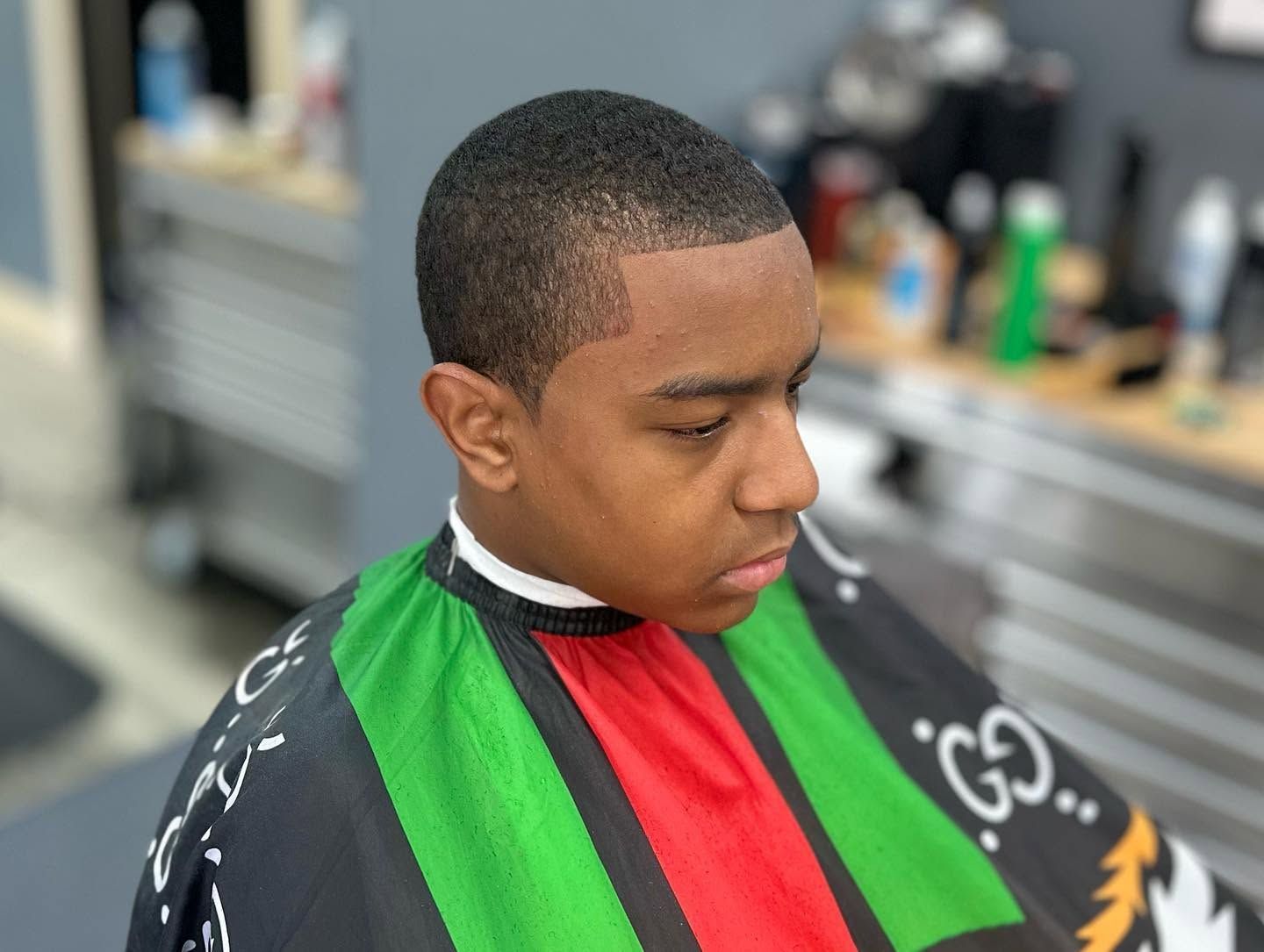 Boy in a barber chair wearing a green, red, and black cape, looking down during a haircut.