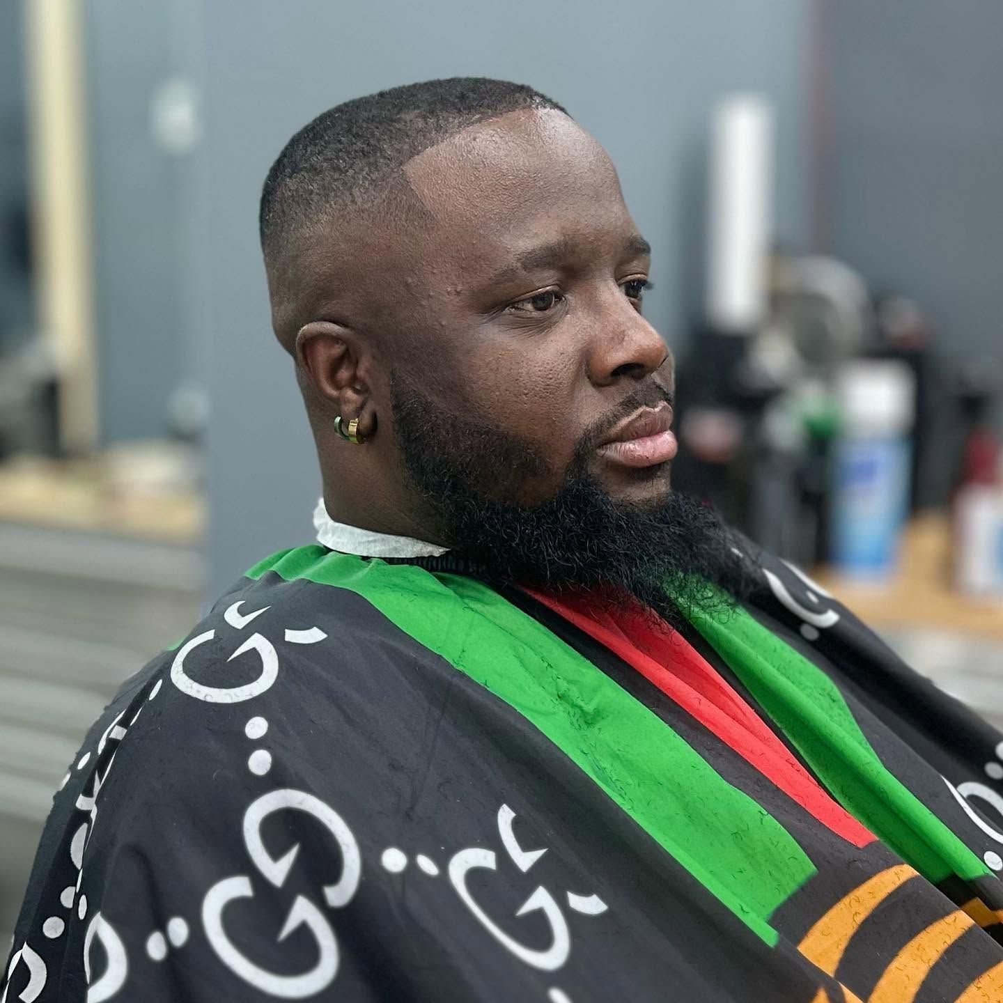 Man seated in a barber chair wearing a black cape with green, red, and gold stripes, looking to the side.