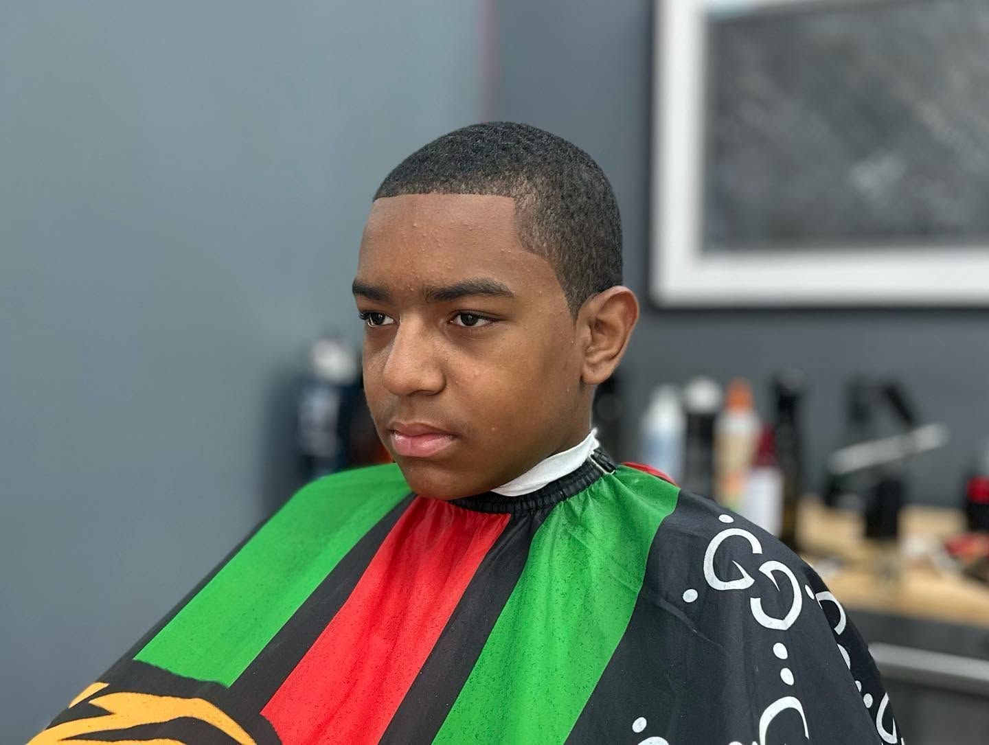 Person in a barber chair wearing a green, red, and black haircut cape, looking serious.