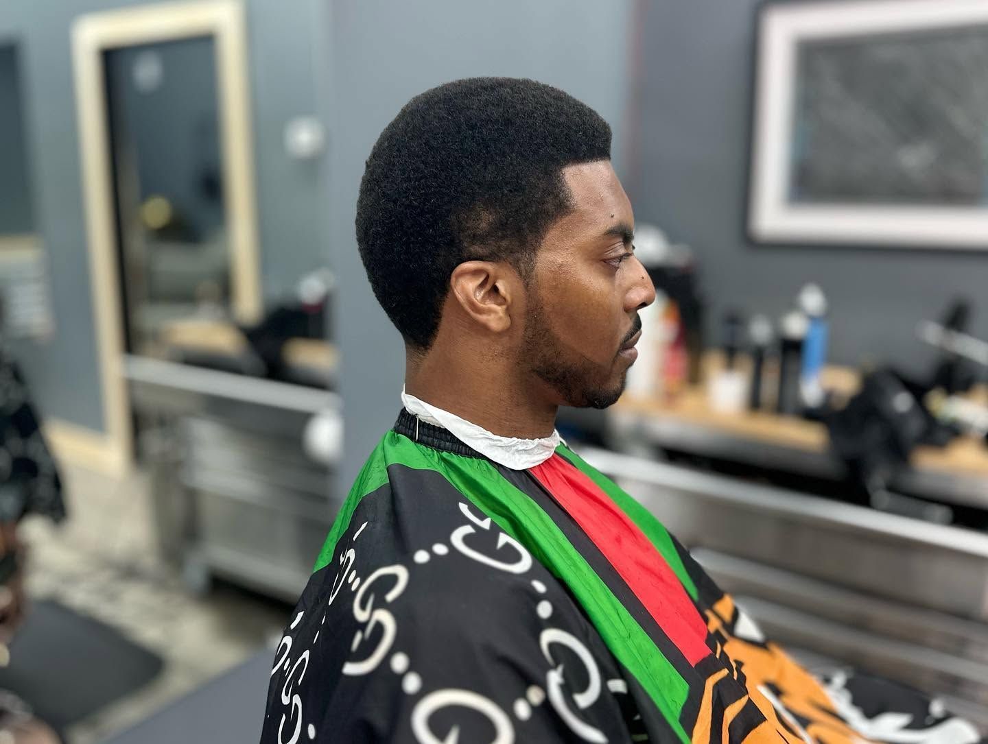 Man seated in a barber chair wearing a black patterned cape with red and green stripes in a barbershop.