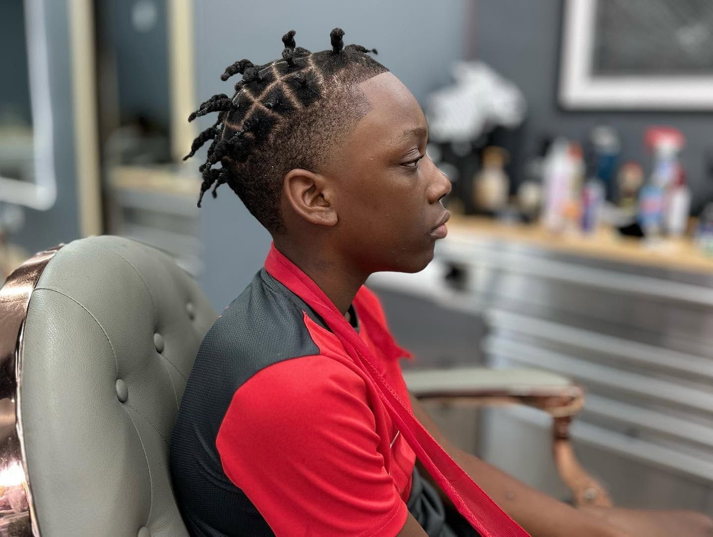 Person with braided hair in a red shirt sitting in a salon chair, viewed in profile.