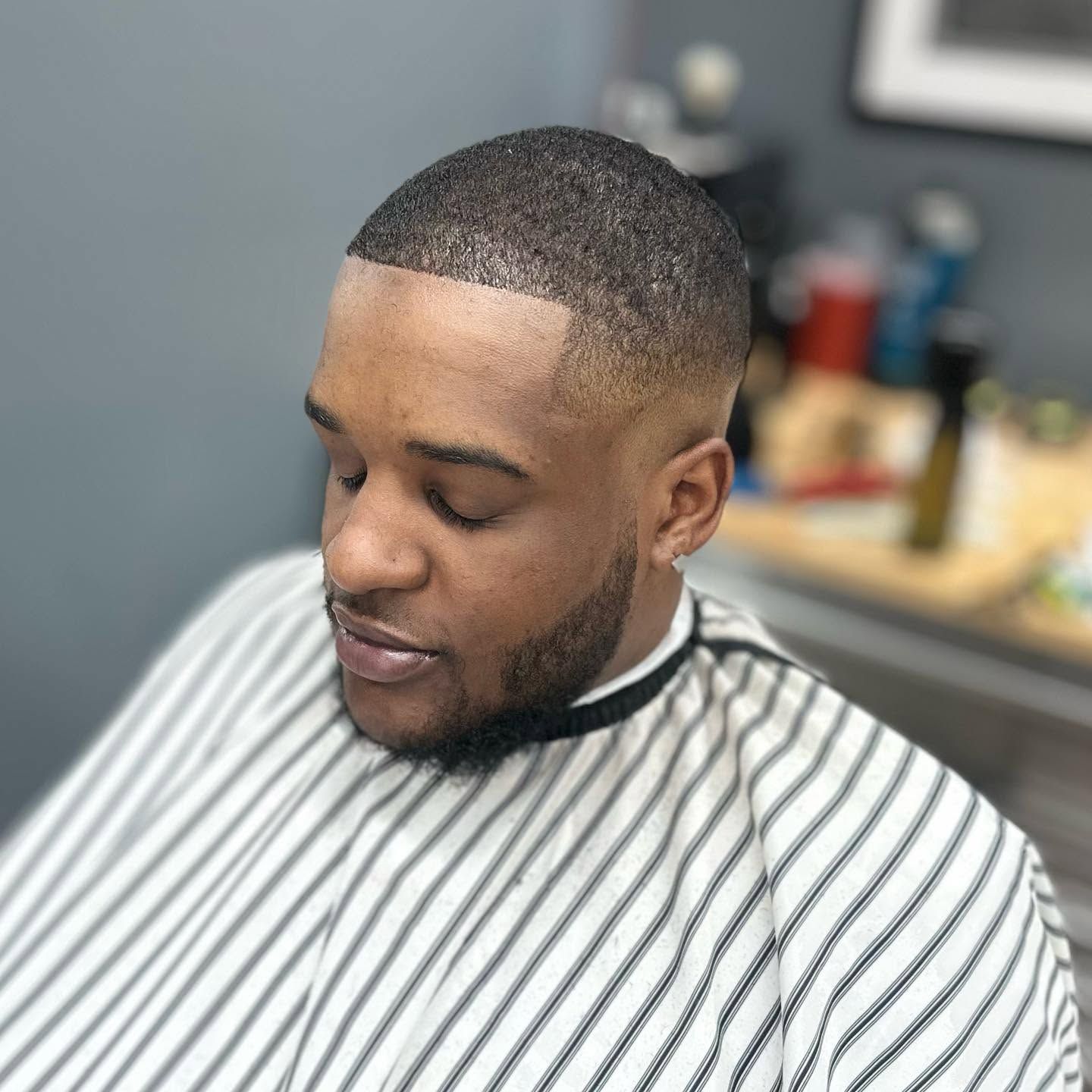 Man with a close-cropped haircut and beard sitting in a barber chair under a striped cape