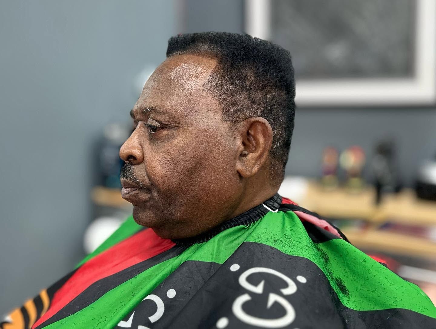 Man seated in a black, green, and red barber cape against a blurred indoor background.