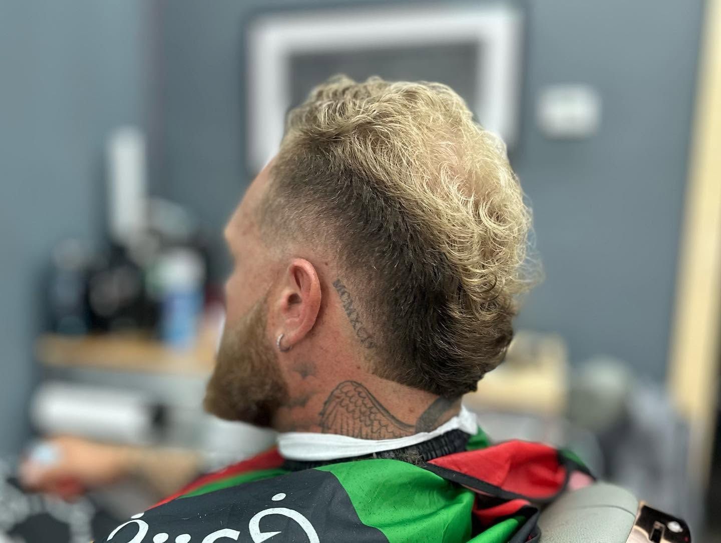 Person with a short blond haircut and neck tattoo seated in a barbershop, wearing a green cape