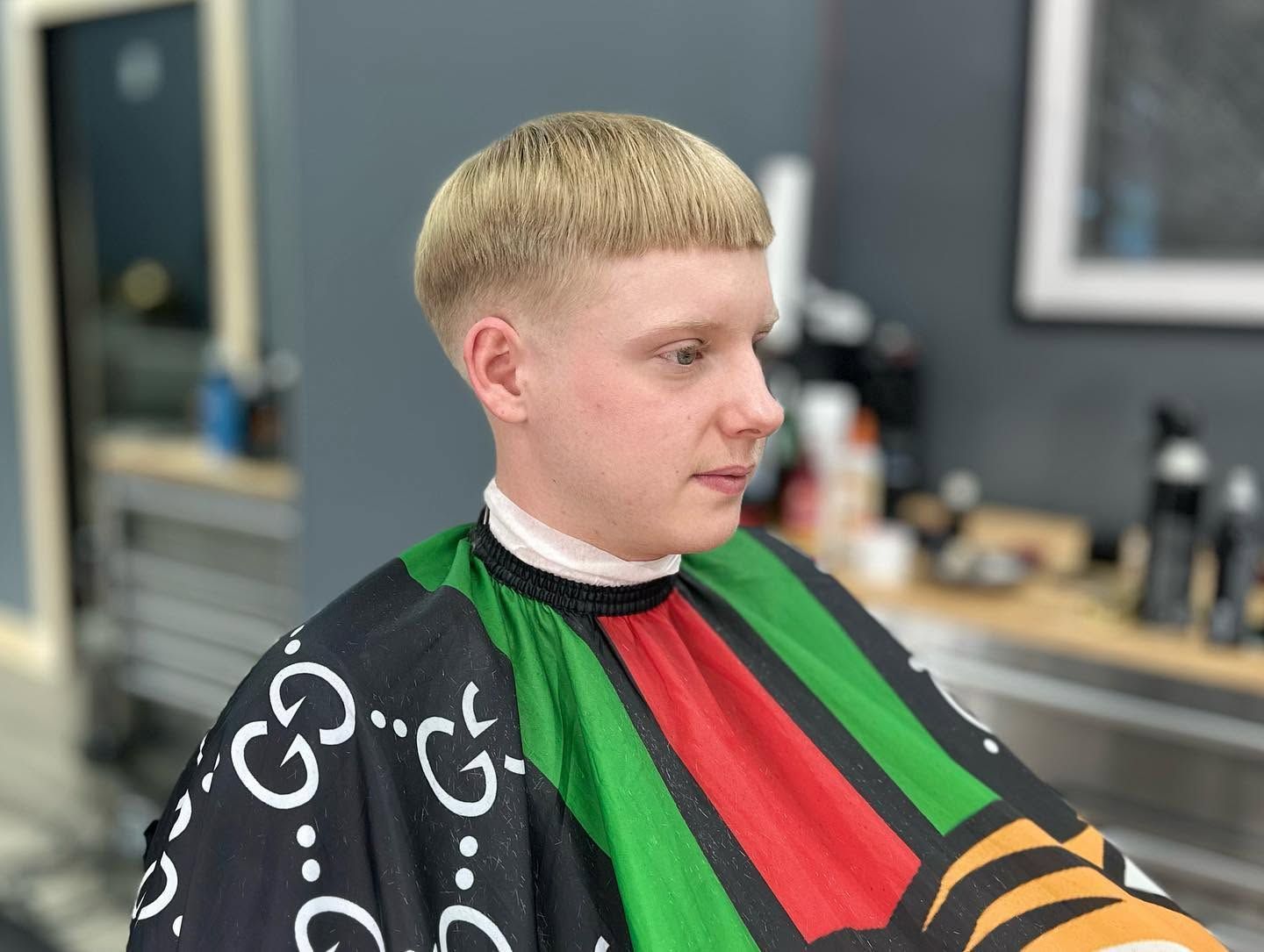 Person with short blond hair wearing a colorful barber cape, seated in a salon chair, looking to the side.