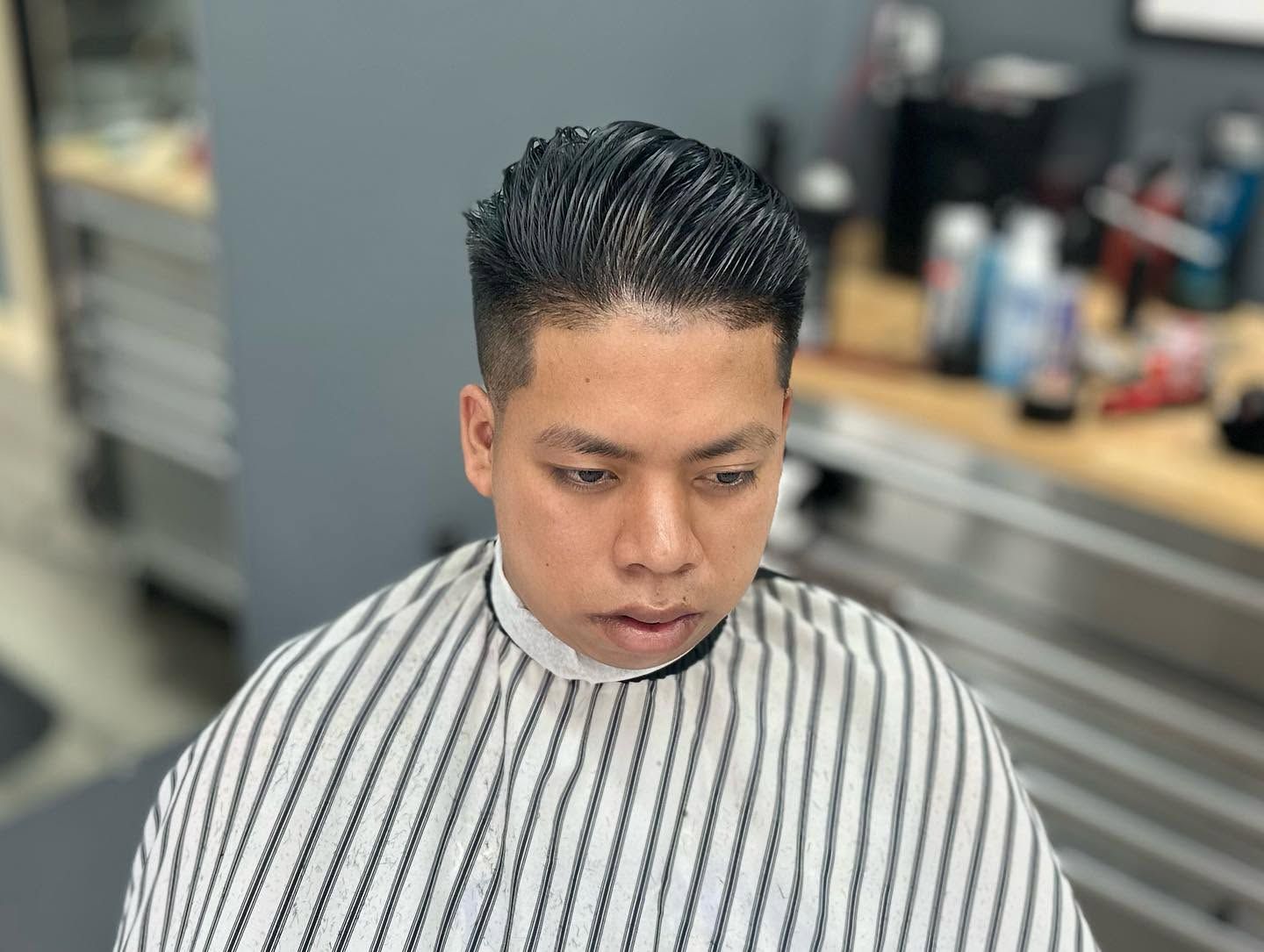 Barber shop customer wearing a striped cape, seated with slicked-back hair, looking down.