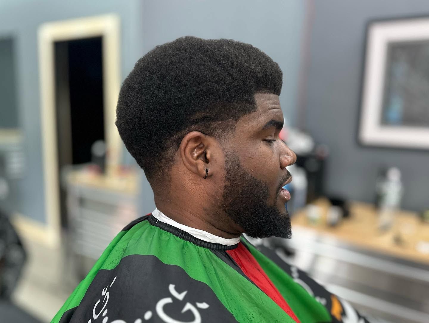 Person in a barber chair wearing a green cape, side profile in a salon with mirrors and tools.