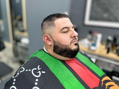 Man getting a haircut in a barbershop, wearing a green cape with red and black stripes, eyes closed.