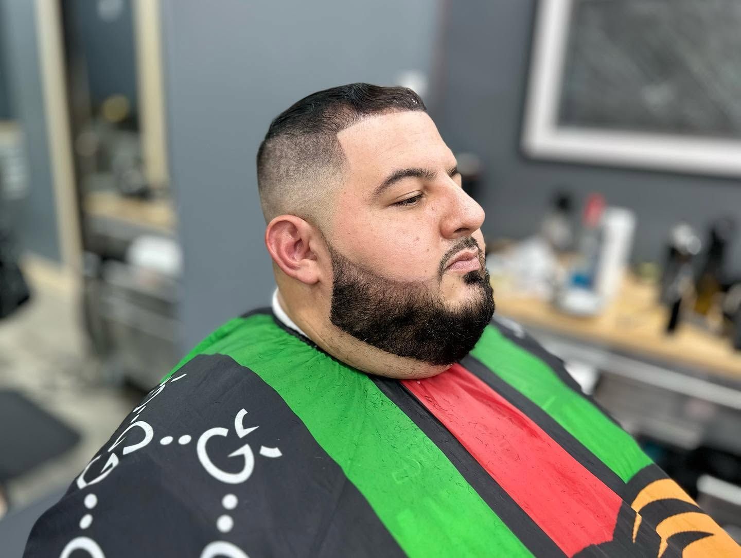 Man with a trimmed beard in a barber chair, wearing a black cape with green and red stripes.
