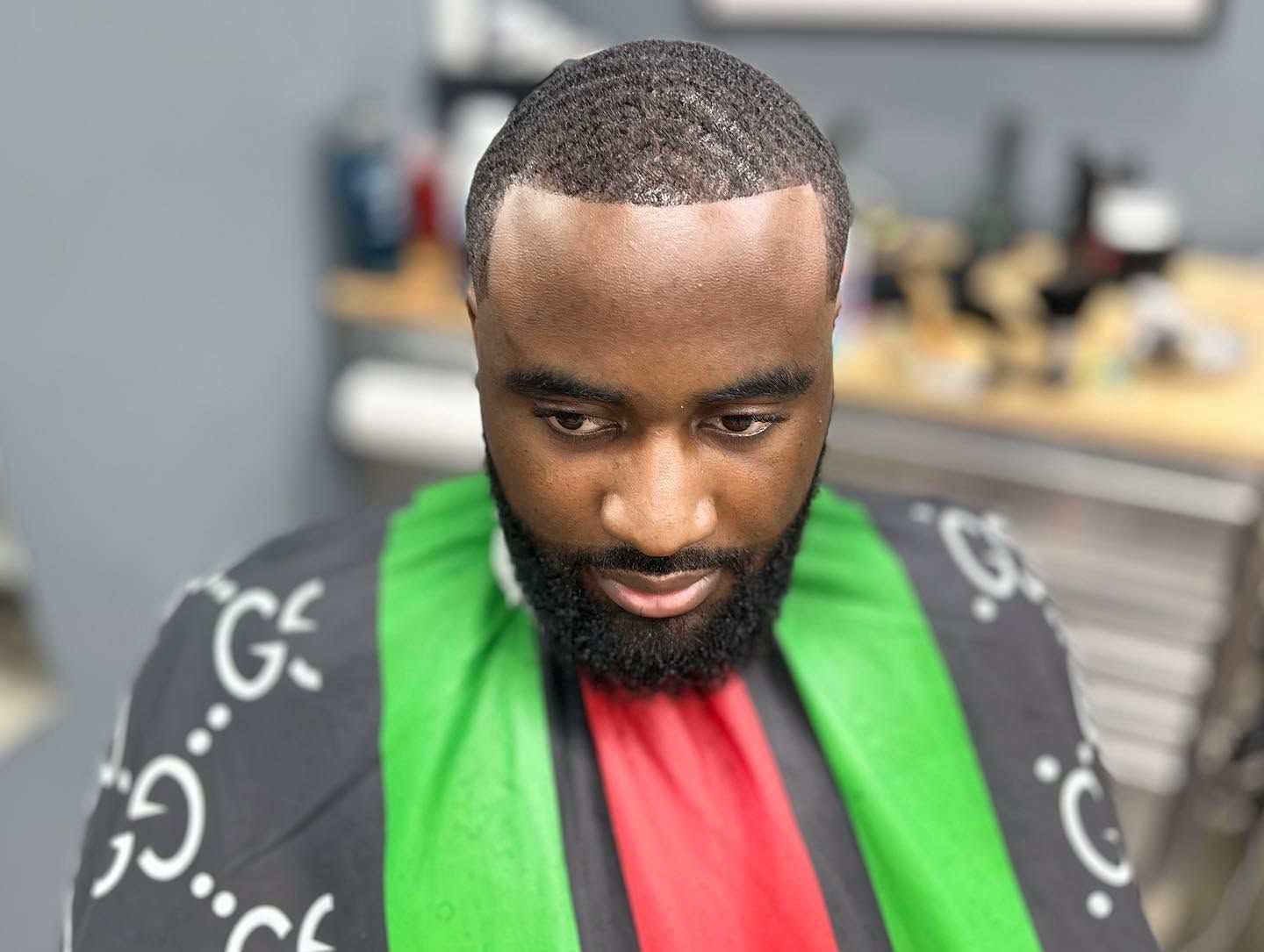 Man with short beard in a barber chair, wearing a green cape with a red strip, in a salon