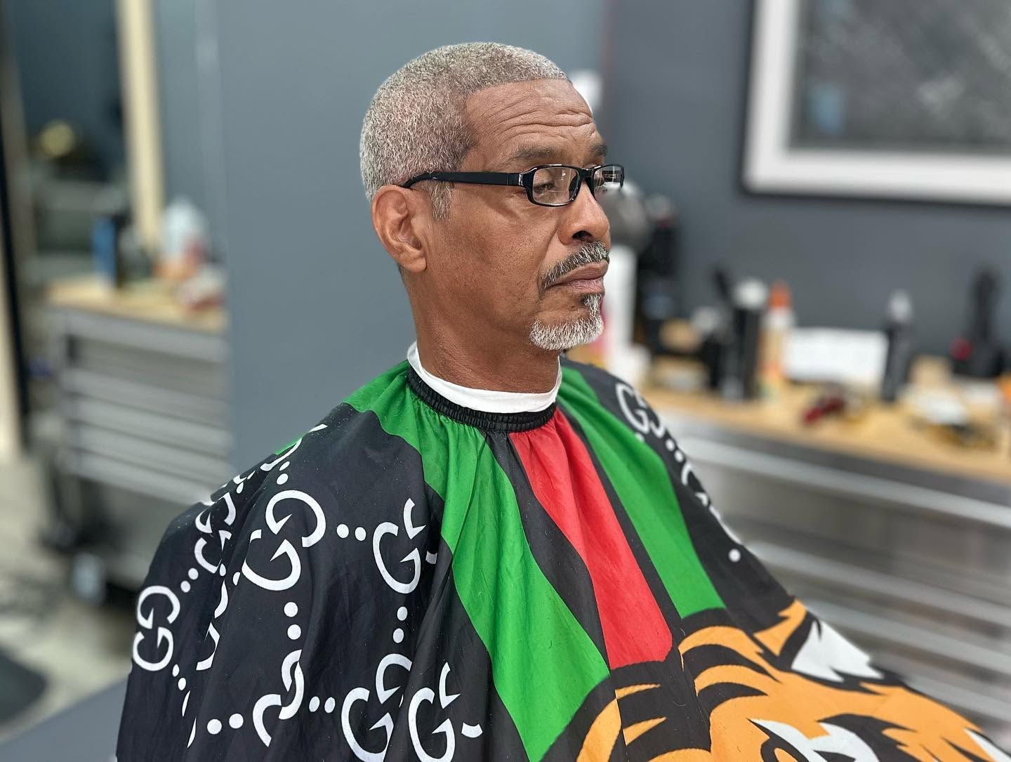 Man wearing glasses and a black-and-green patterned shirt in a barbershop, seated and looking to the side