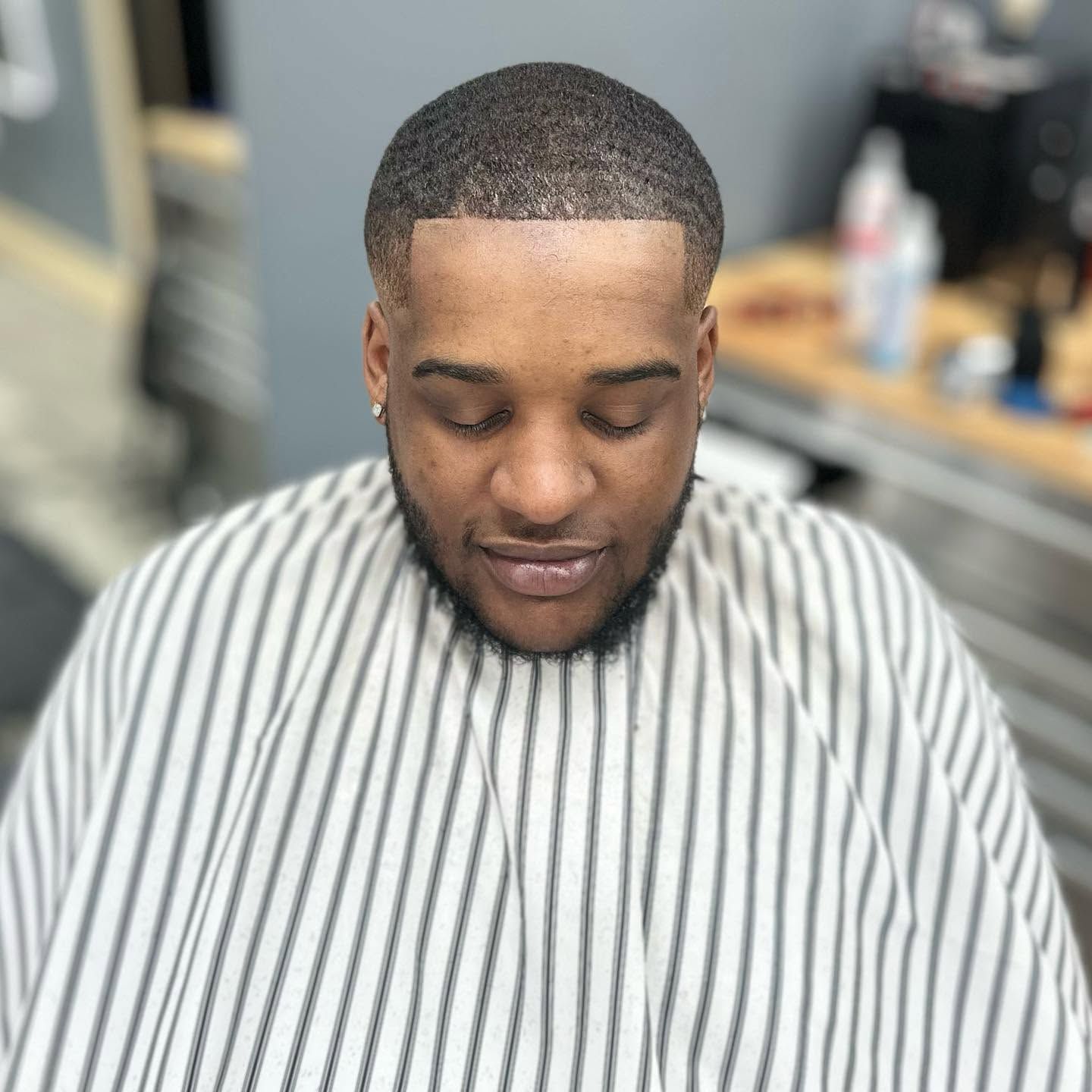 Man getting a close-cropped haircut in a barber chair, wearing a striped cape.