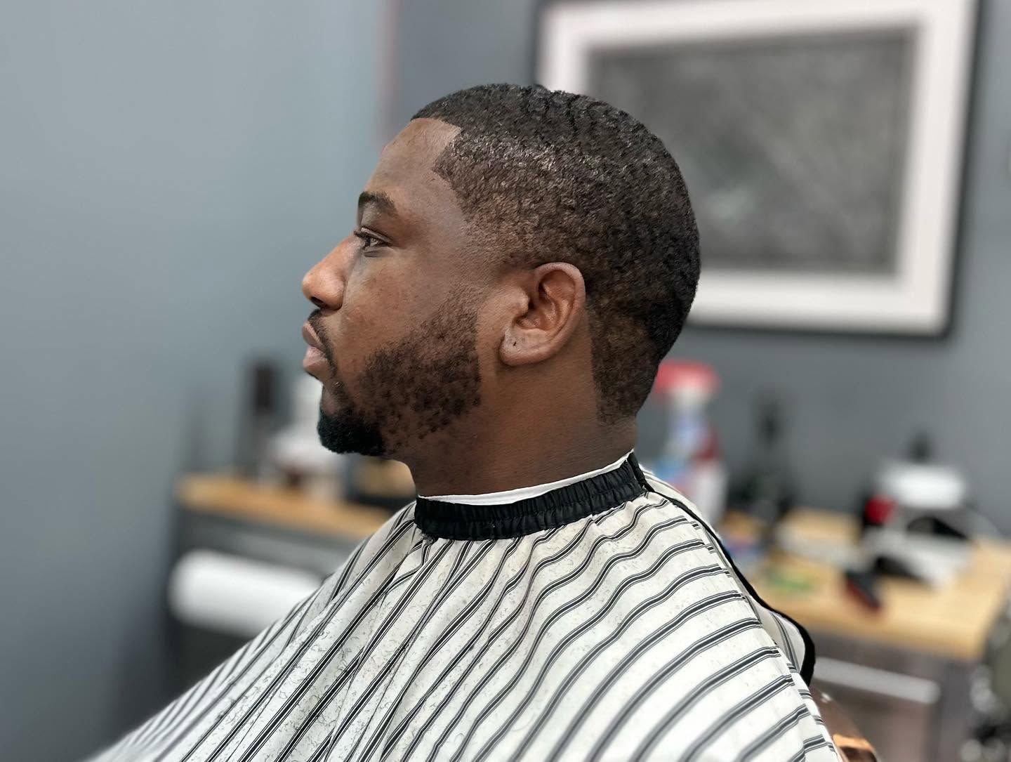 Man in a barber chair wearing a striped cape, shown in profile inside a barbershop.