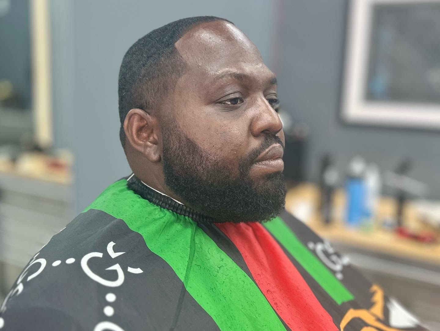 Man with trimmed beard wearing a green, red, and black barber cape, seated in a salon chair