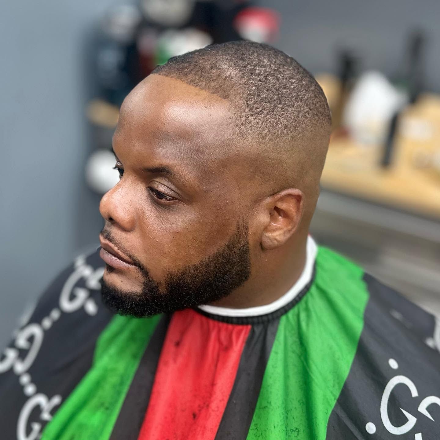 Side-profile of a man with a short haircut and beard wearing a black, green, and red barber cape.