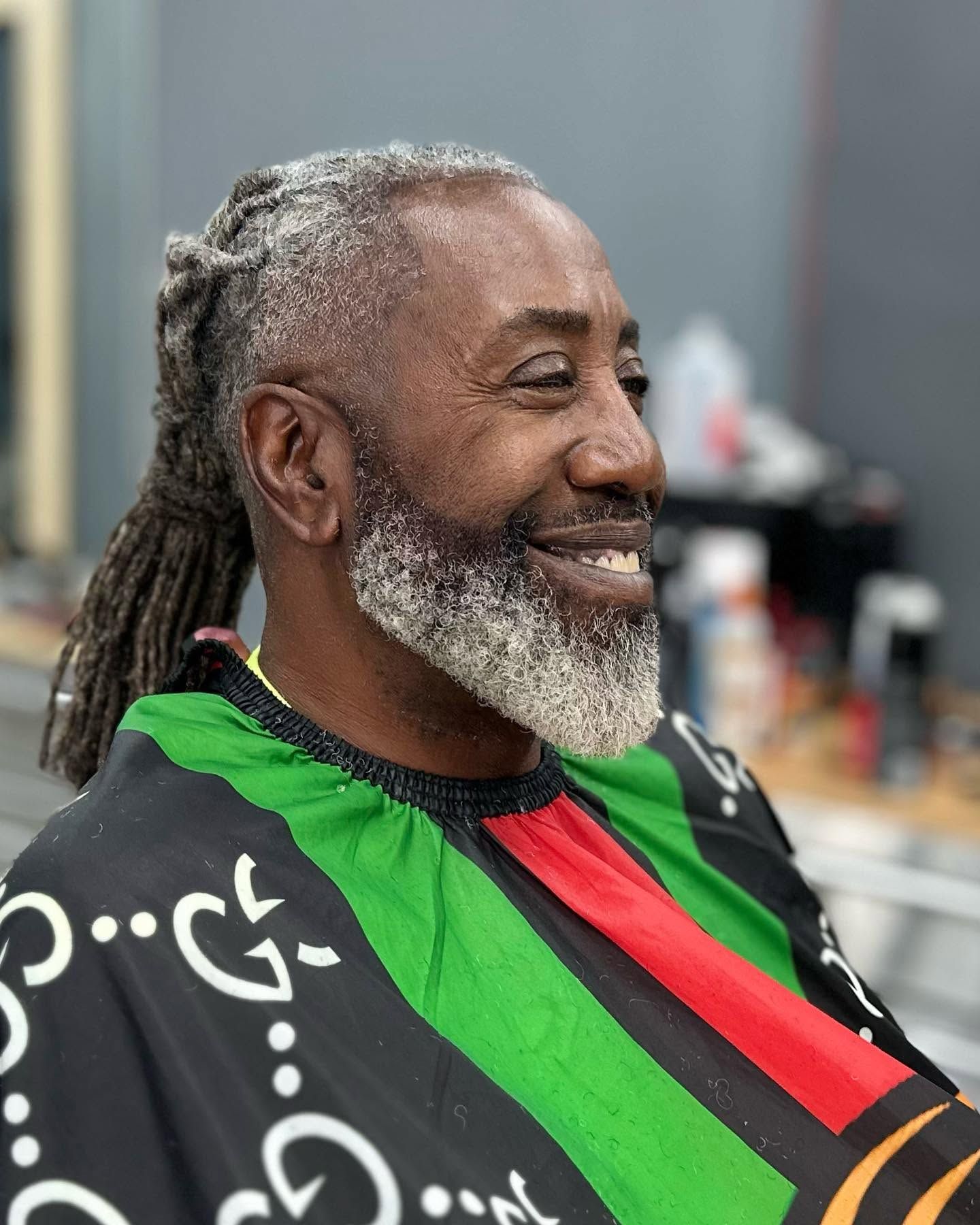Smiling man with a gray beard wearing a black, green, and red barber cape in a salon chair