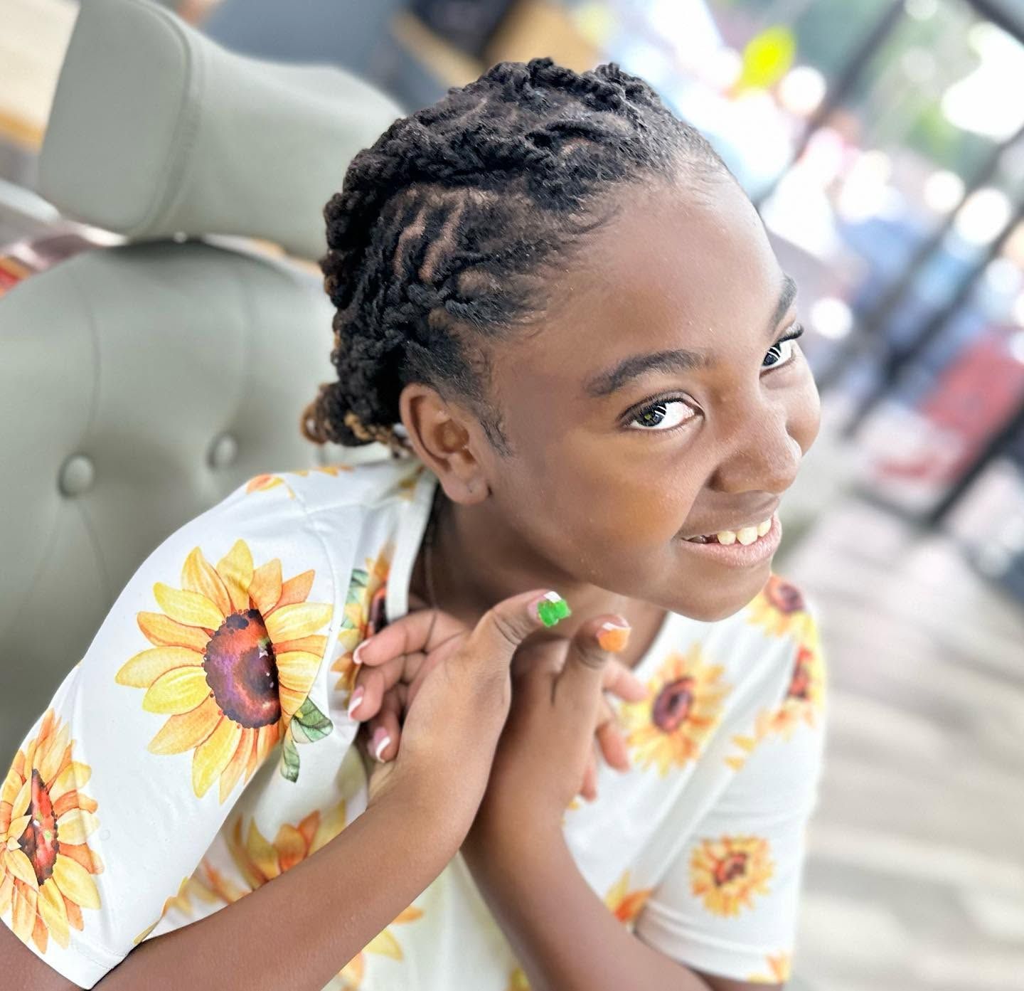 Smiling child with braided hair in a sunflower shirt, hands clasped, sitting on a chair outdoors.