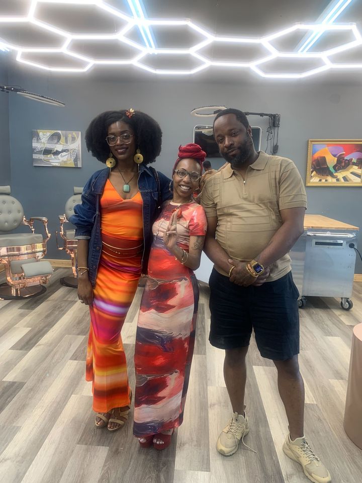 Three people posing indoors under geometric ceiling lights, with colorful outfits and framed art in the background.