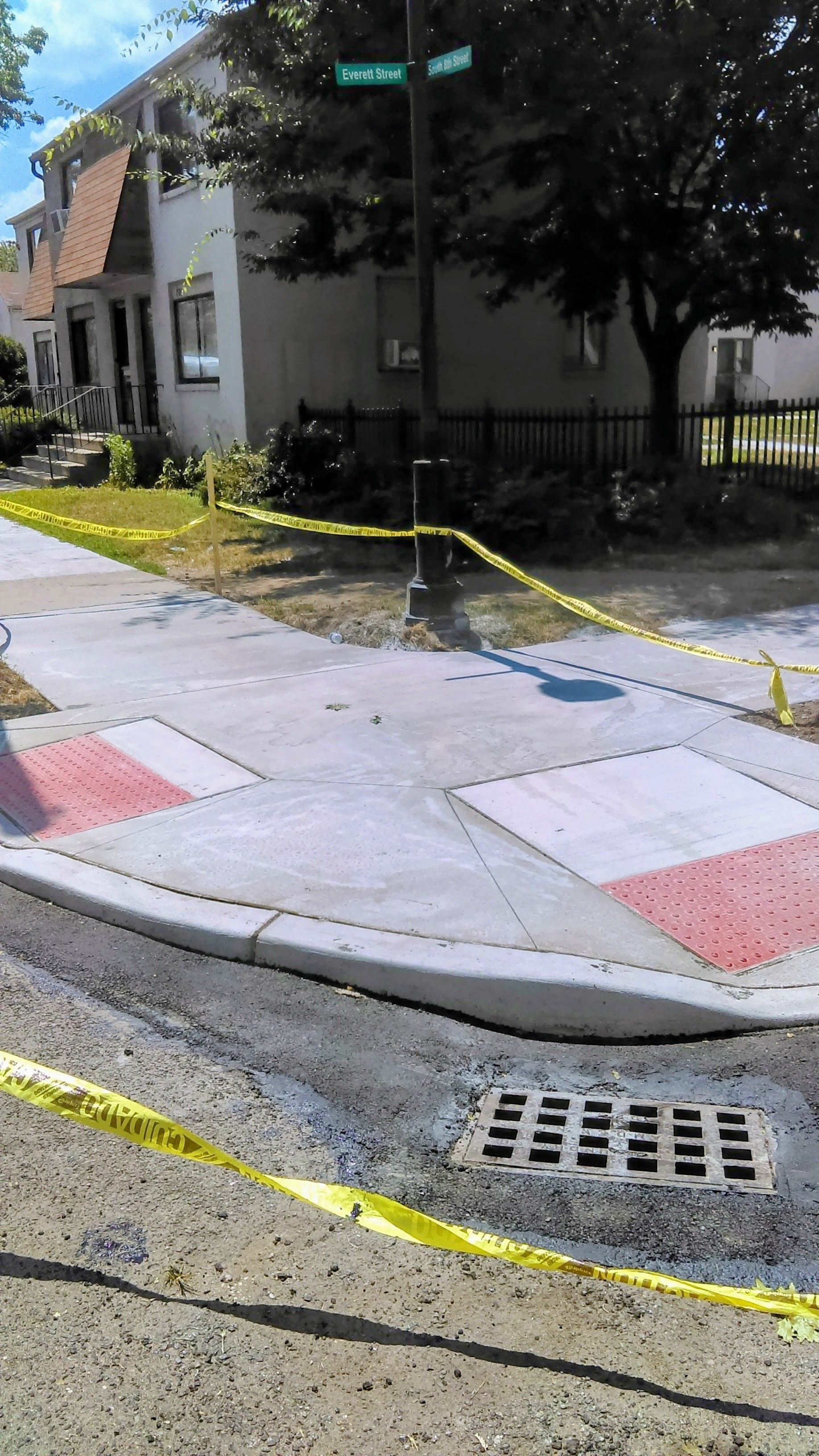 New sidewalk construction at a street corner with detectable warning surface, caution tape, and a drain.
