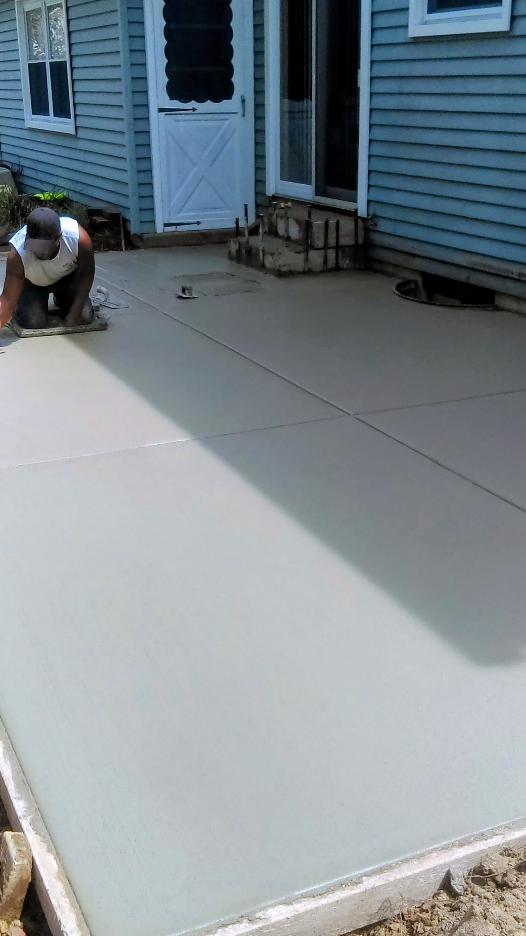 Person smoothing fresh concrete on a patio outside a blue house.