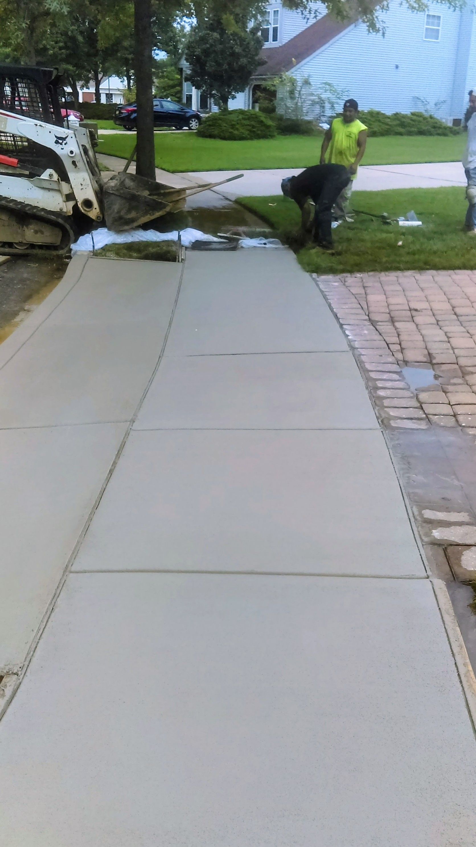 Workers pouring concrete for a driveway. A Bobcat and wheelbarrow are also present on a sunny day.