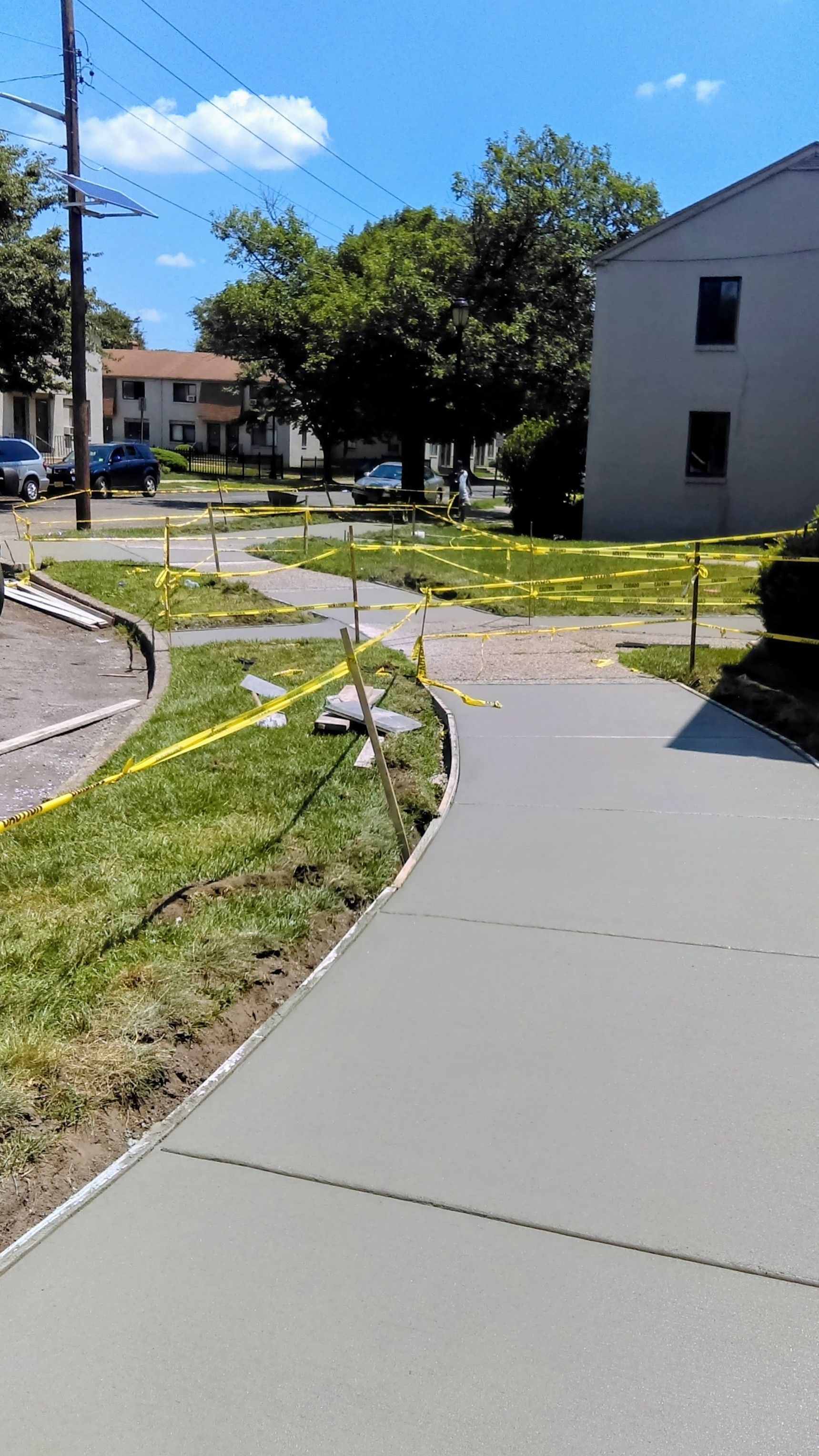 Newly poured concrete sidewalk with caution tape, adjacent to grass, under a blue sky.