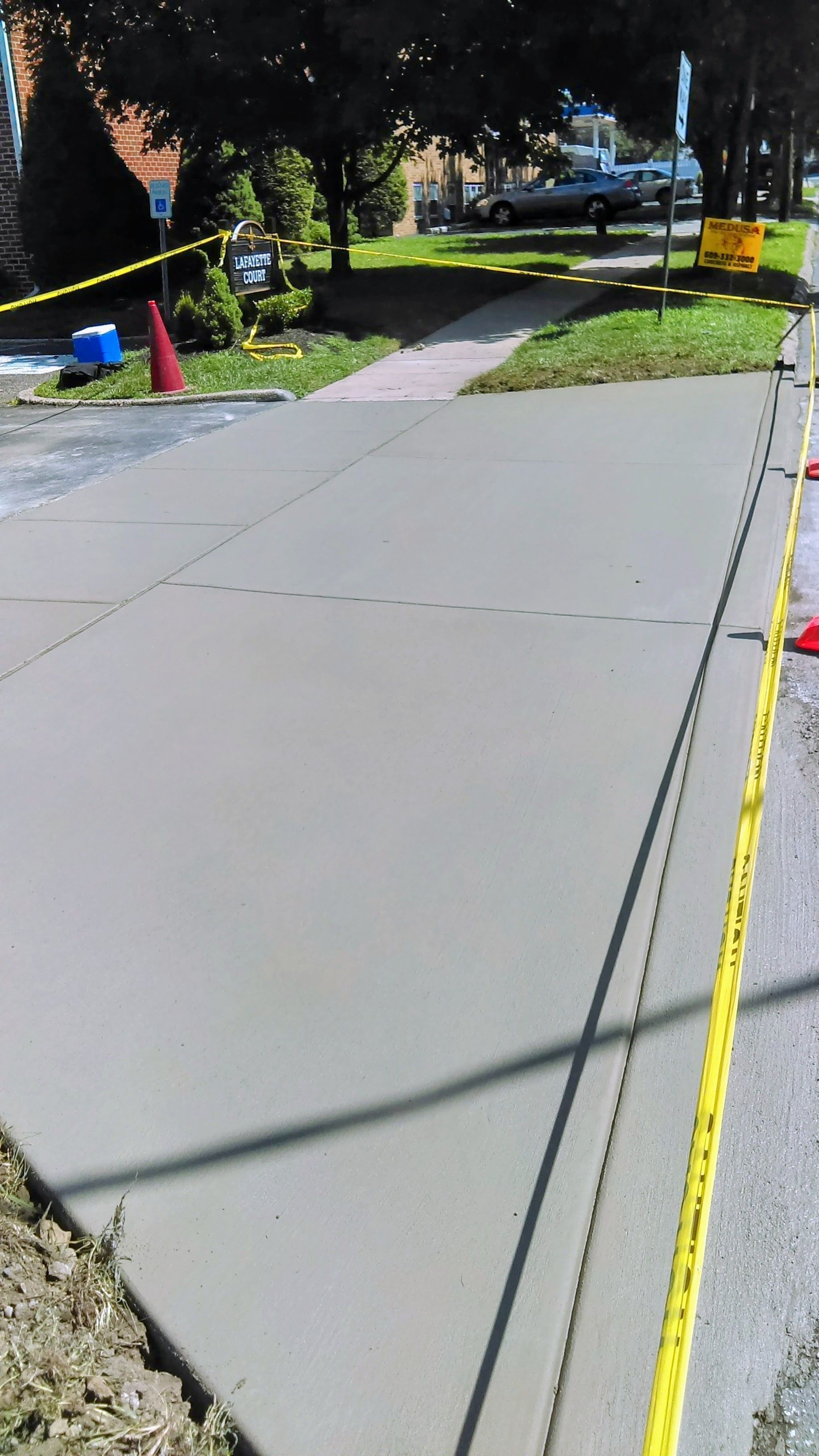 Freshly poured concrete sidewalk with yellow caution tape, cones, and a residential street in the background.