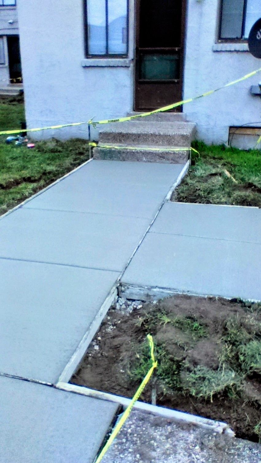 Newly poured concrete walkway and steps leading to a building entrance, bordered by grass and caution tape.