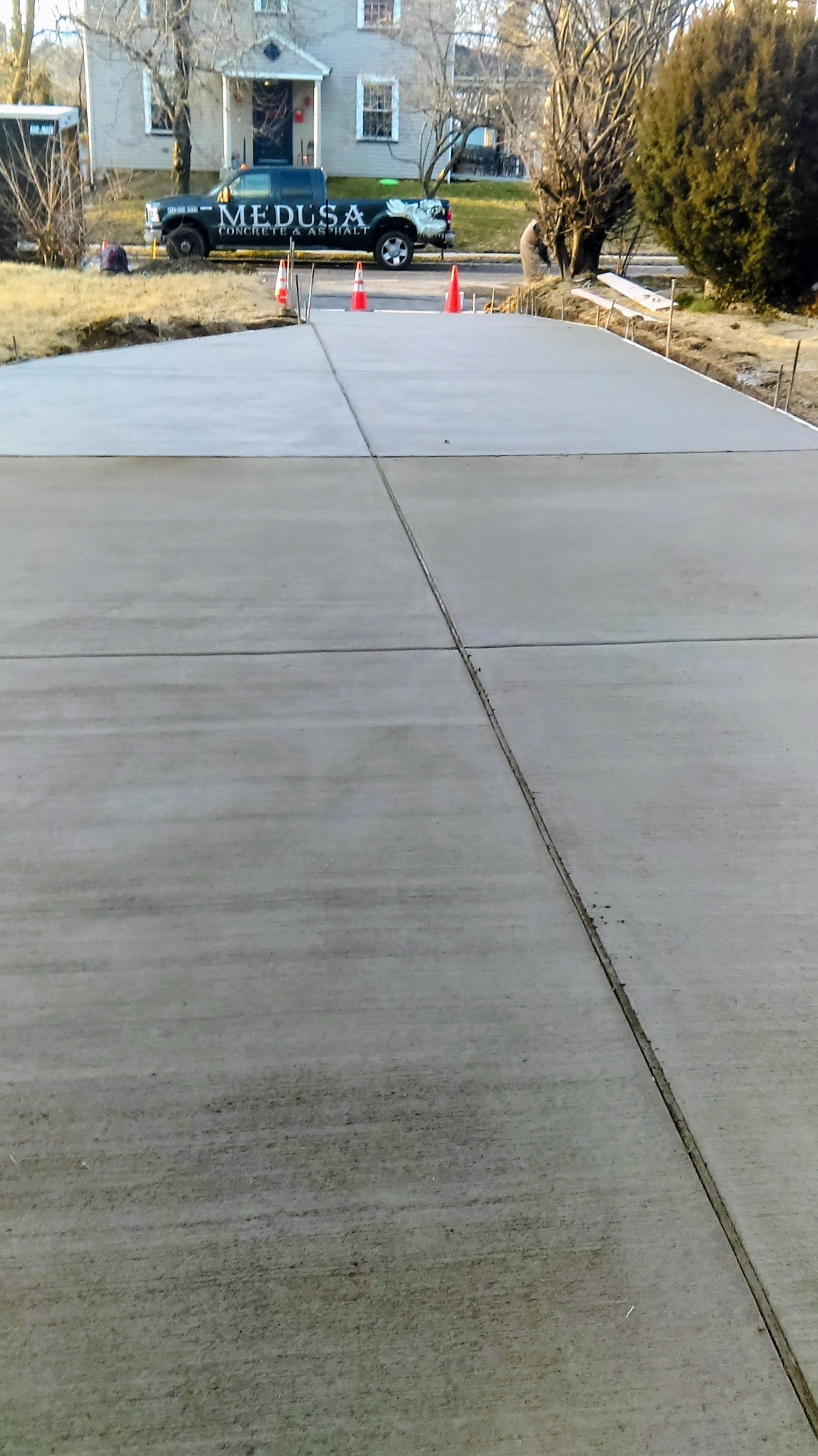 Newly poured concrete driveway with a truck in the background, surrounded by orange traffic cones.