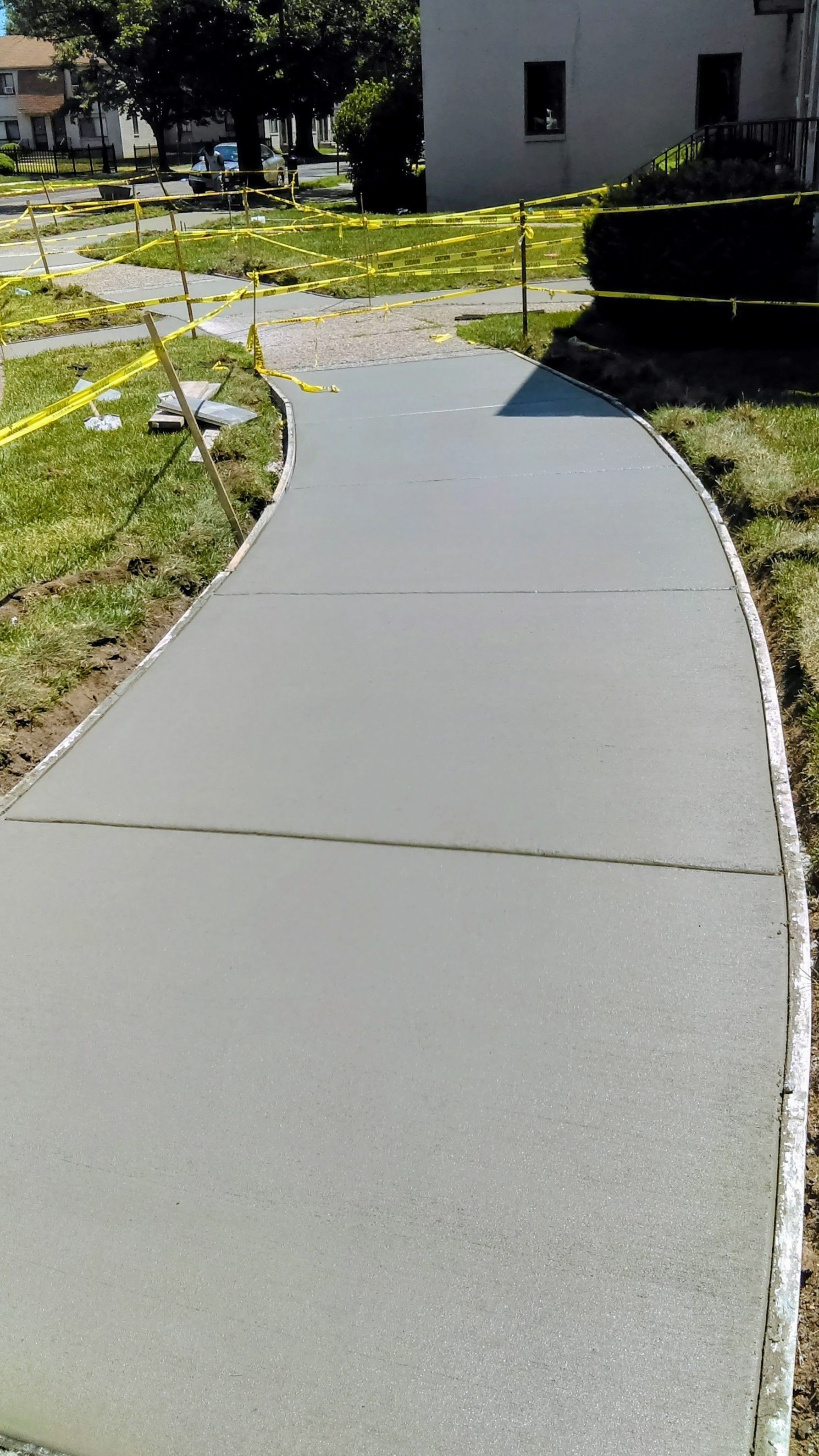Newly poured concrete sidewalk curves through a grassy yard, cordoned off with yellow caution tape.