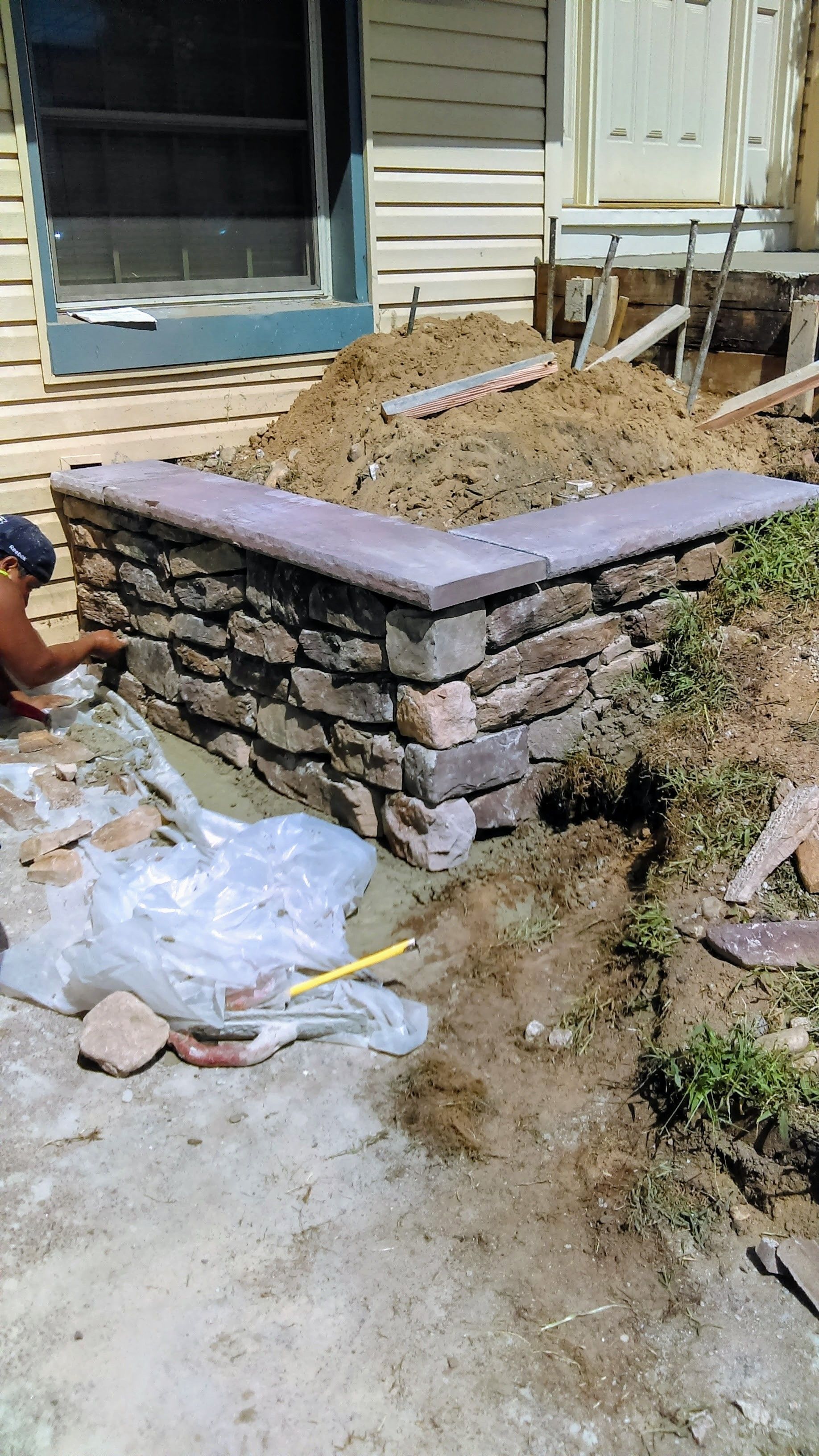 A brick retaining wall being built near a house. Construction materials, dirt, and a person are present.