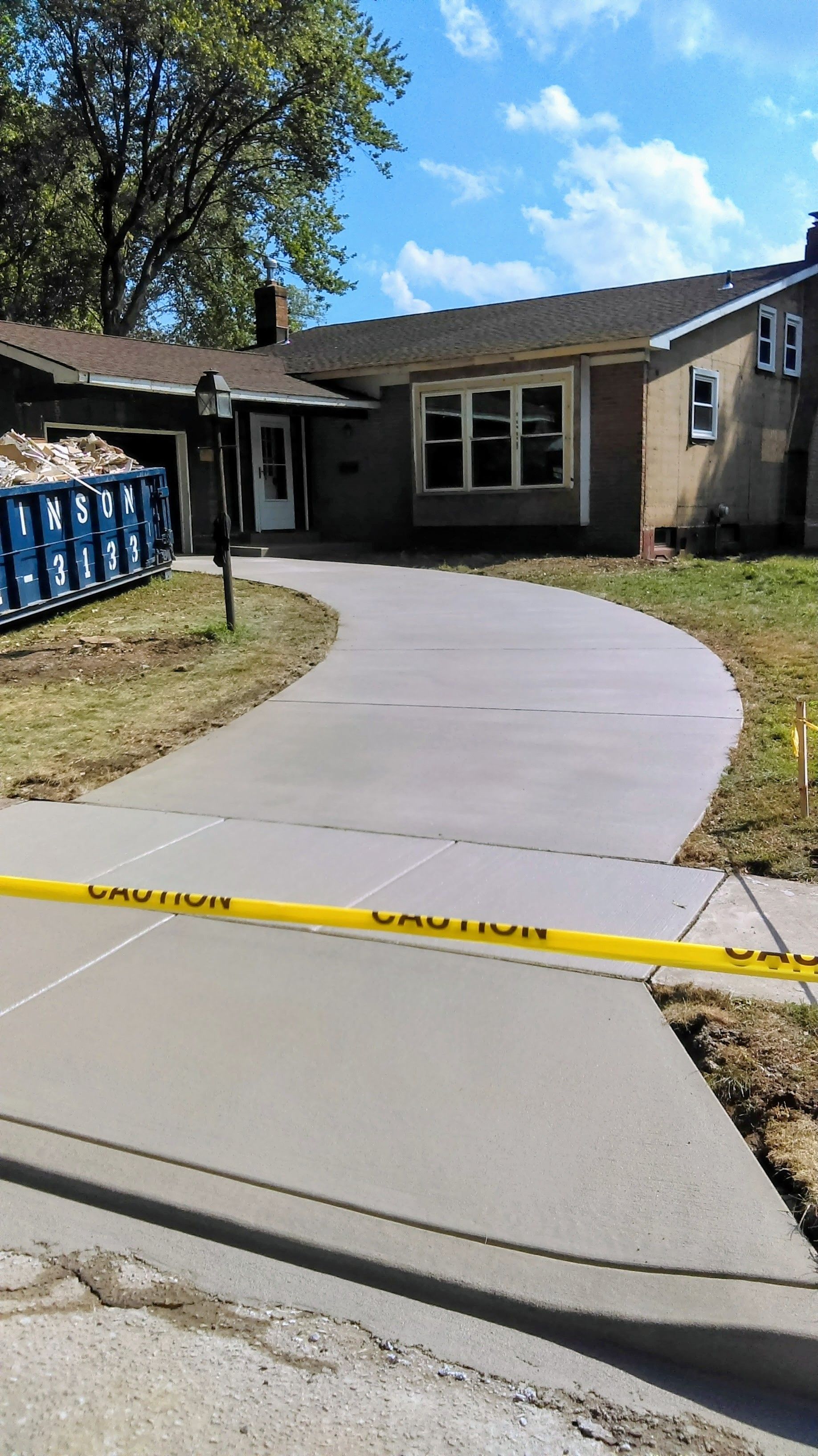 Newly poured concrete driveway curves toward a house, yellow caution tape across it.
