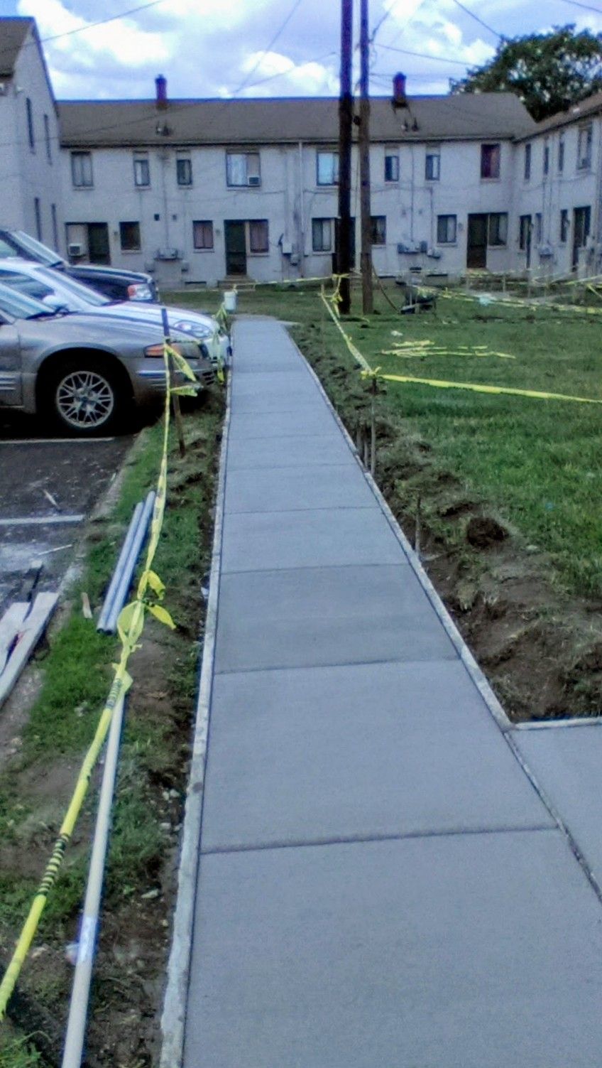 Newly poured concrete sidewalk next to grass and parked cars, with a row of apartments in the background.