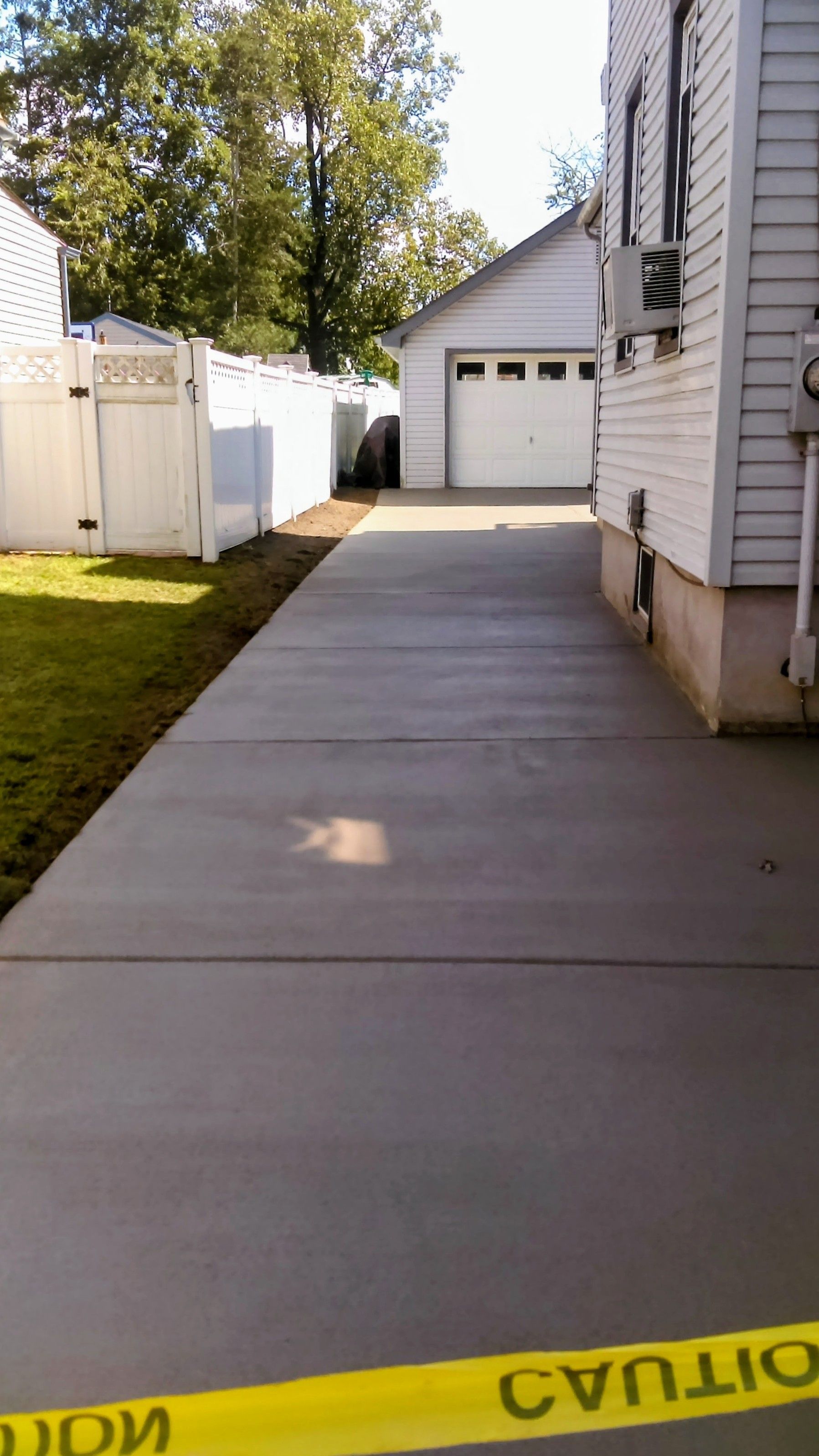 Concrete driveway leads to a garage.  White fence and house on either side. Yellow caution tape in foreground.
