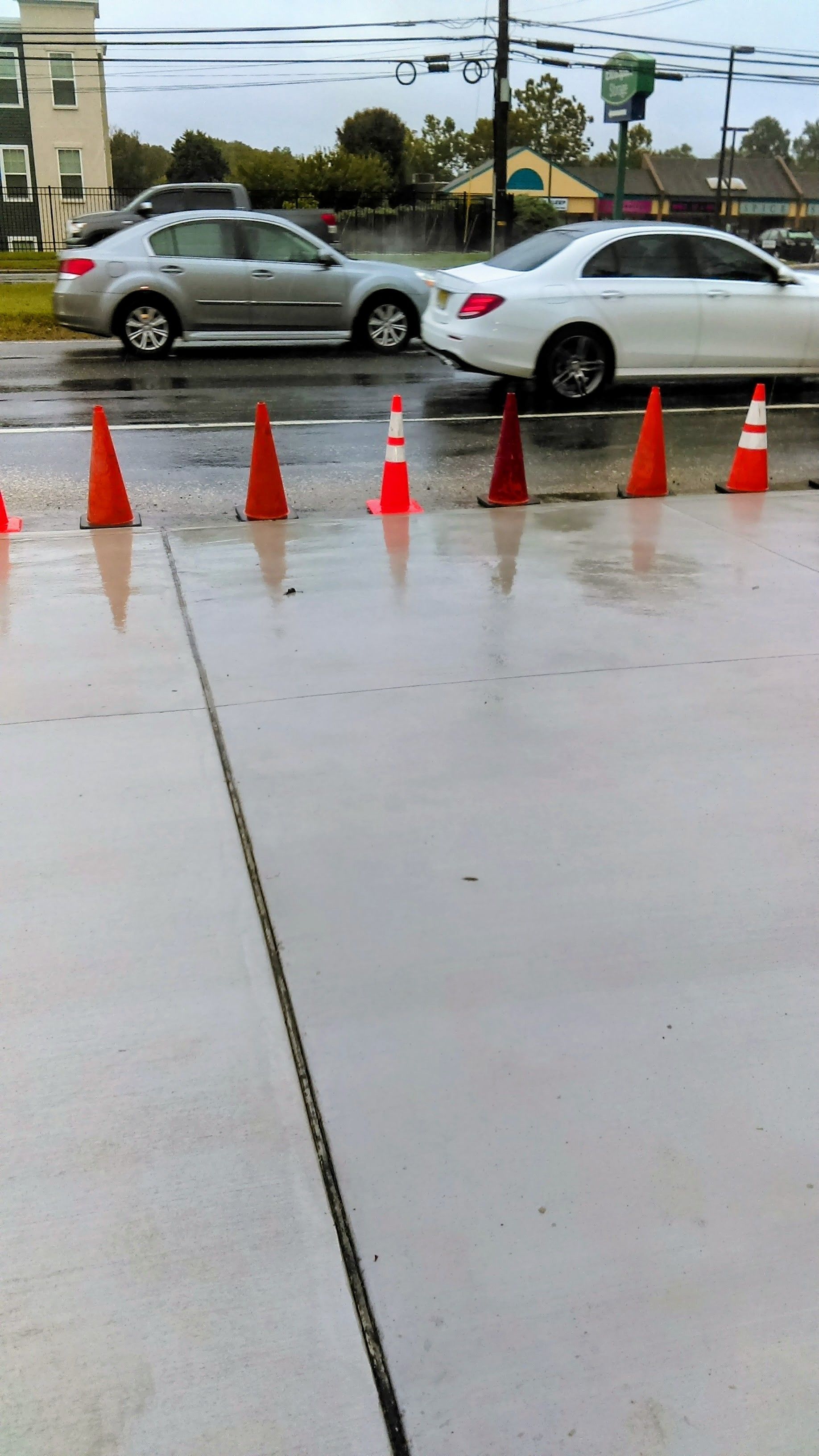 Wet road with orange cones, silver and white cars, overcast sky.