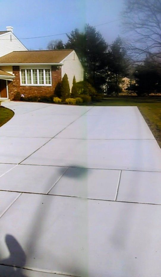Driveway in front of a brick house with a brown roof; green lawn and trees in the background under a blue sky.