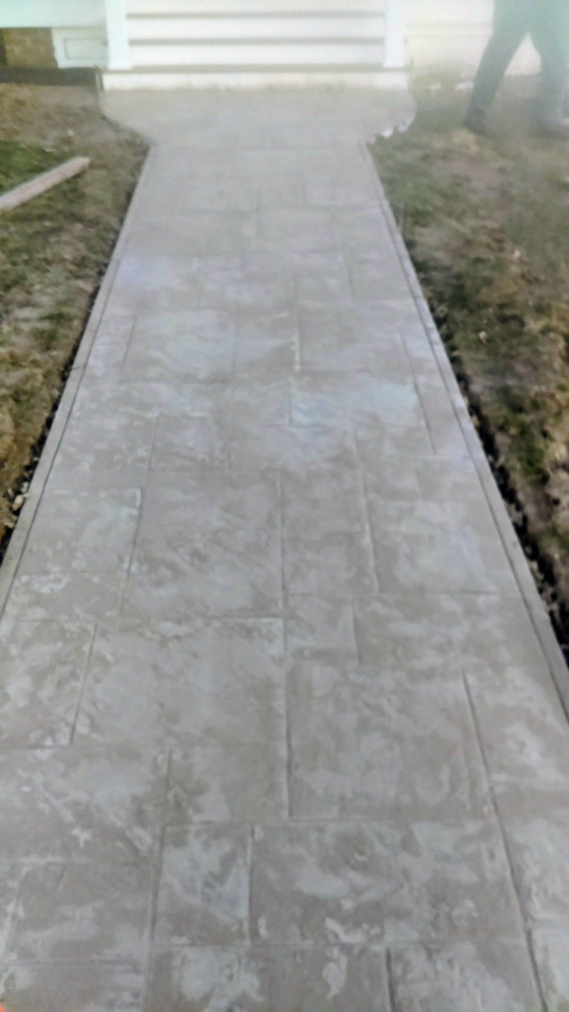 Concrete walkway leading to a building, light gray with rectangular tile pattern. Grass and a person's legs are visible.