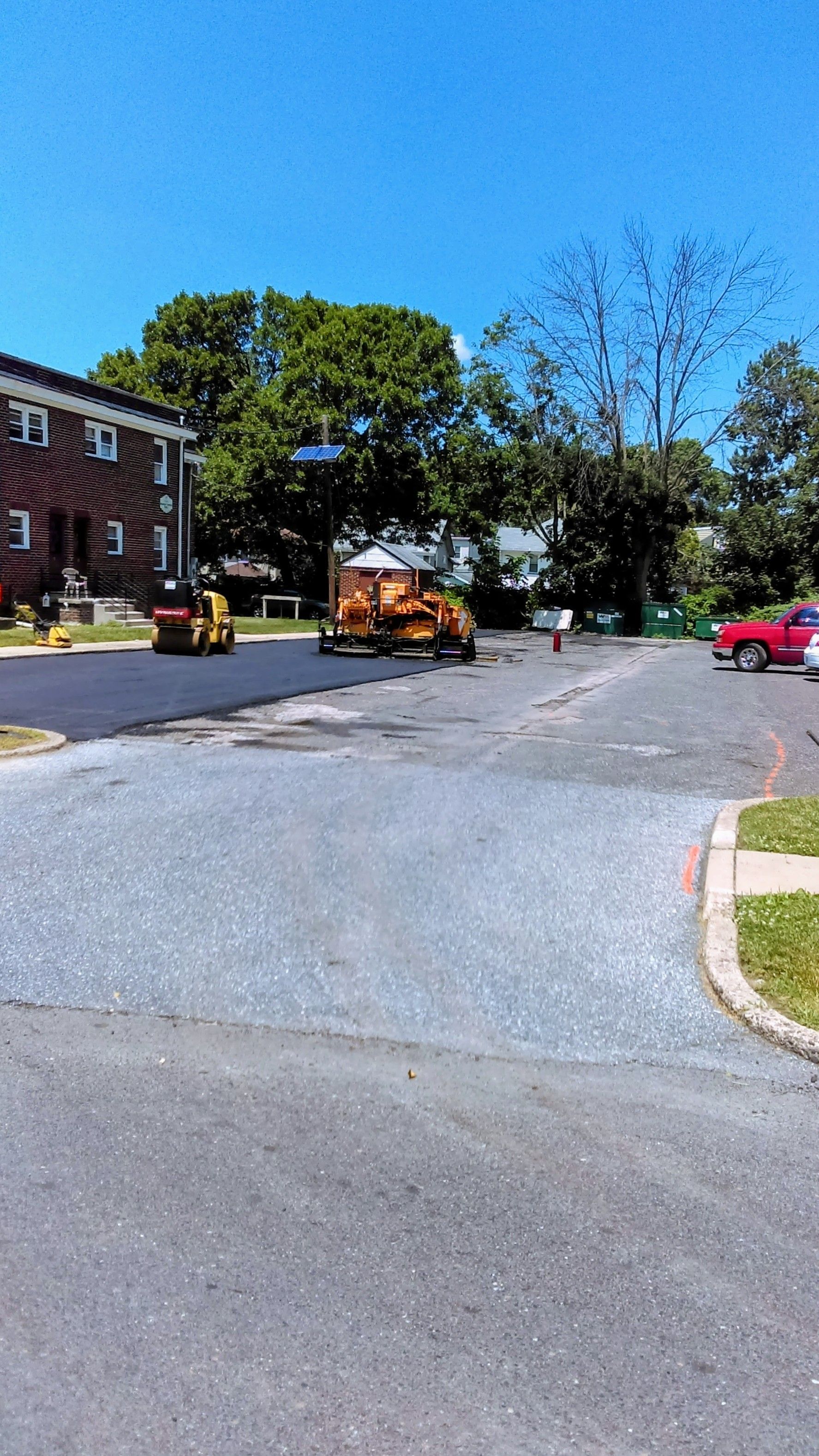 Road construction site with machinery; blue sky, trees, and buildings visible.