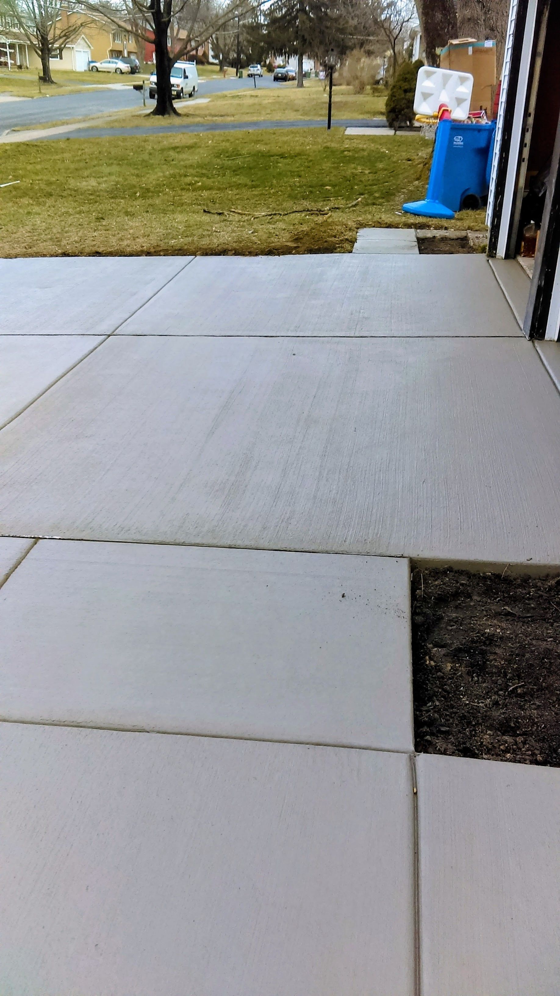 Concrete walkway with missing square, green grass, and blue trash can in the background.