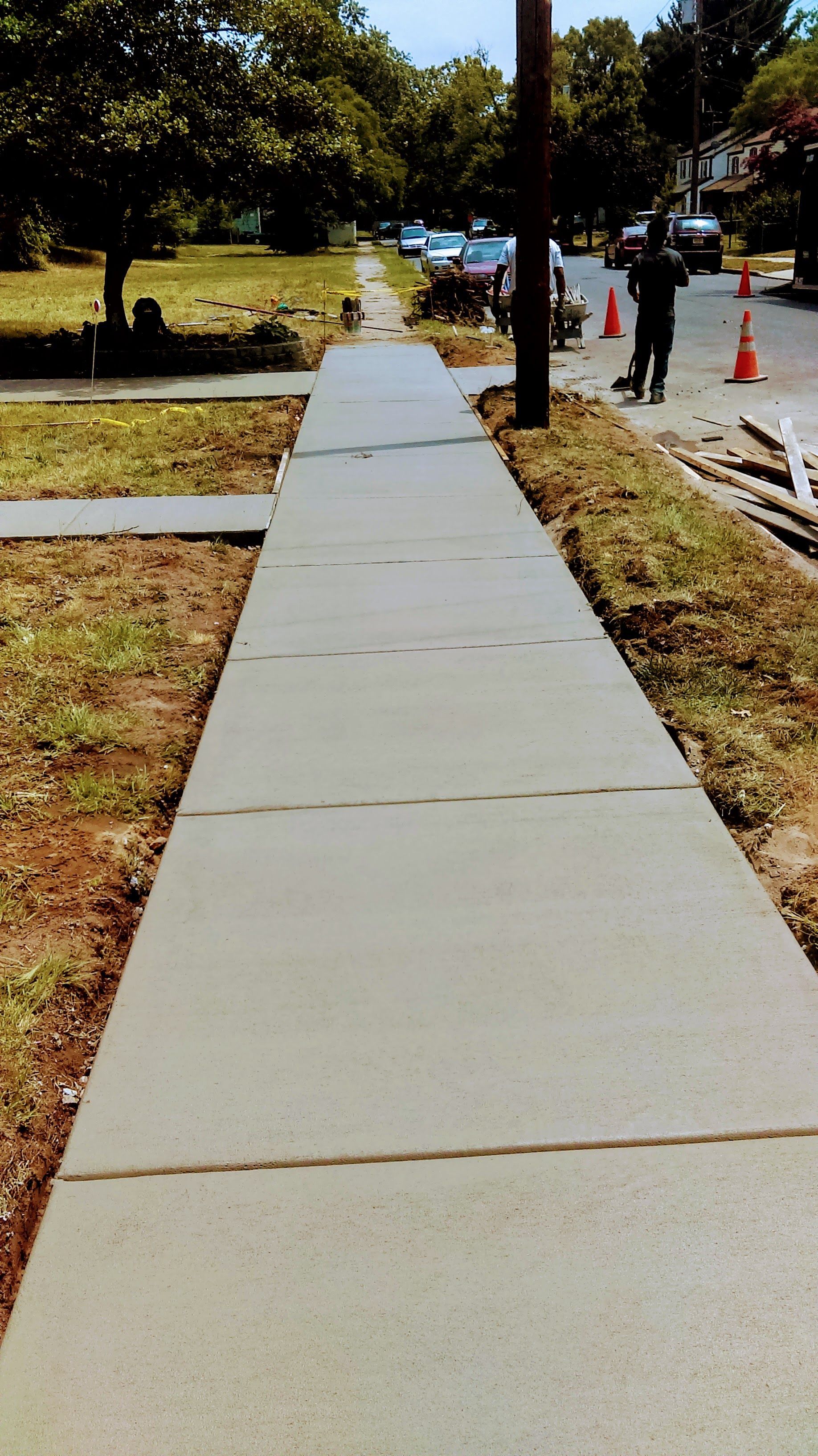Freshly poured concrete sidewalk next to a street with cars, construction cones, and a worker.