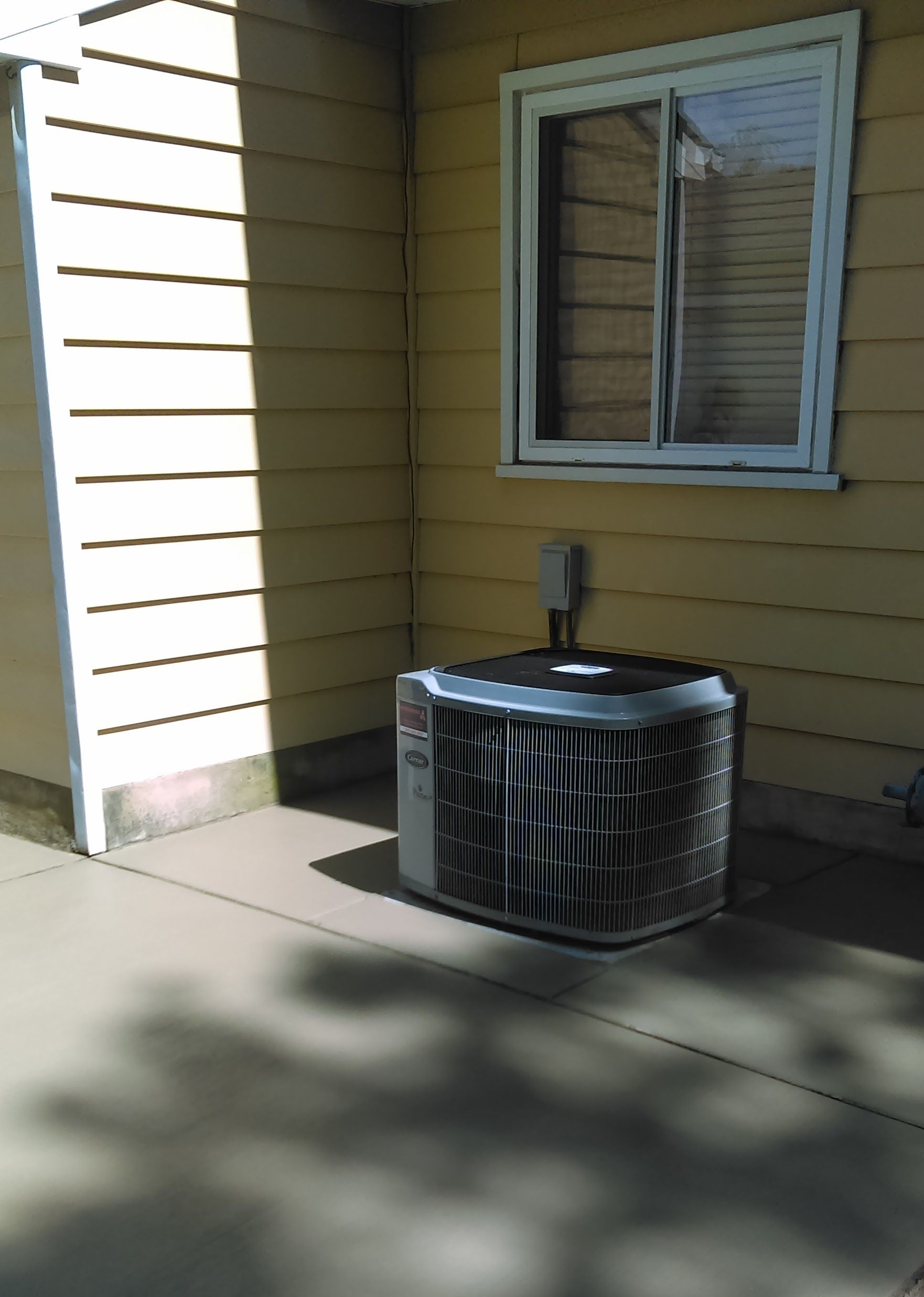 Exterior air conditioning unit next to a house with yellow siding and a window.