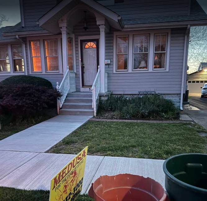A light blue house with a gray cement path and lawn. Sign on the left, trashcan on the right.