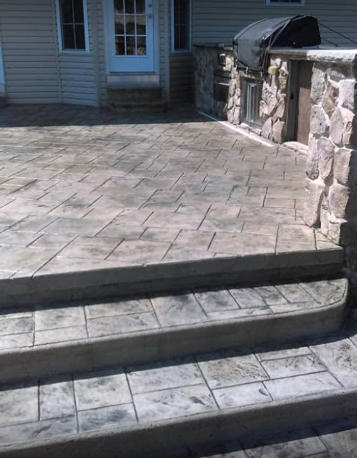 Stamped concrete patio with steps leading to a house entrance. Stone grill and walls are on the right.