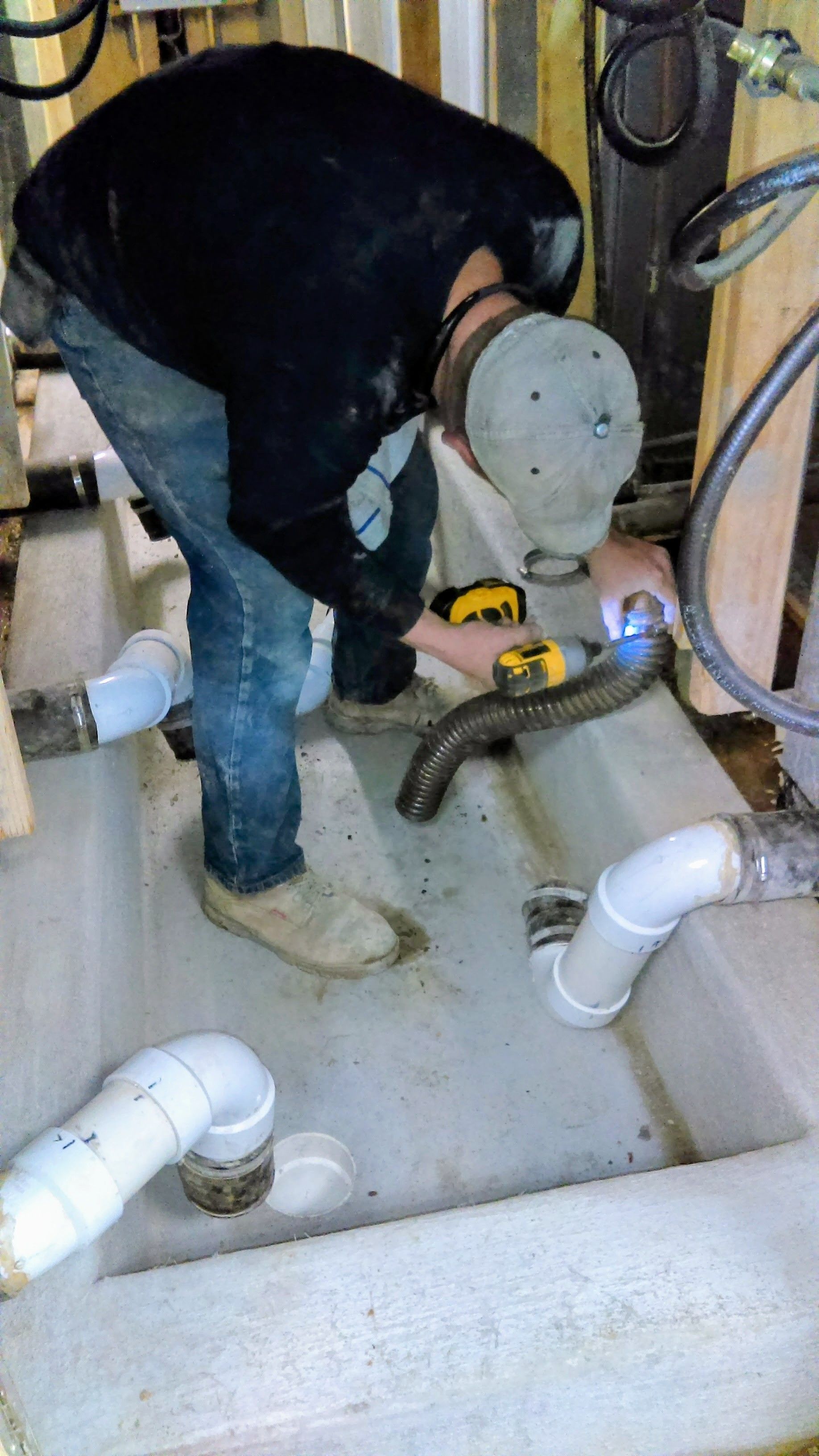 Person using a power tool to work on pipes in a concrete trough; wearing jeans, a black jacket, and a hat.