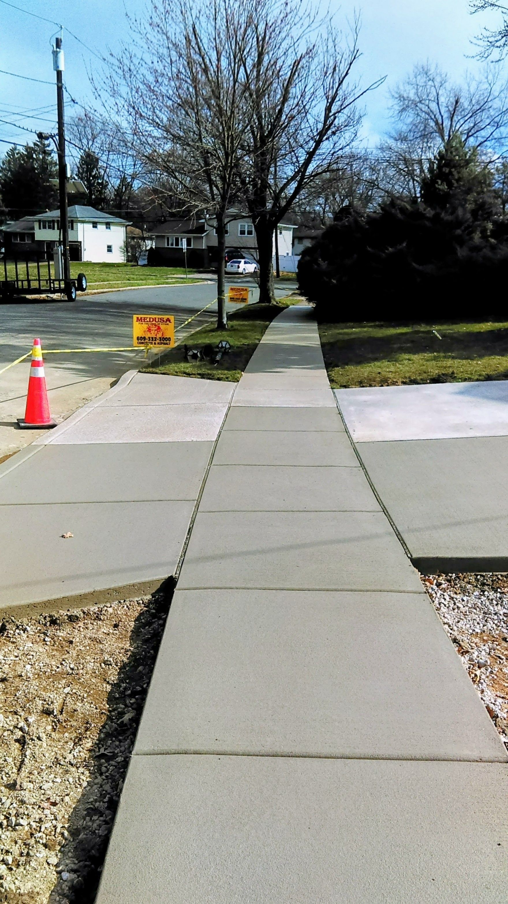 Concrete sidewalk leading toward a tree, houses in the distance, and construction signs.