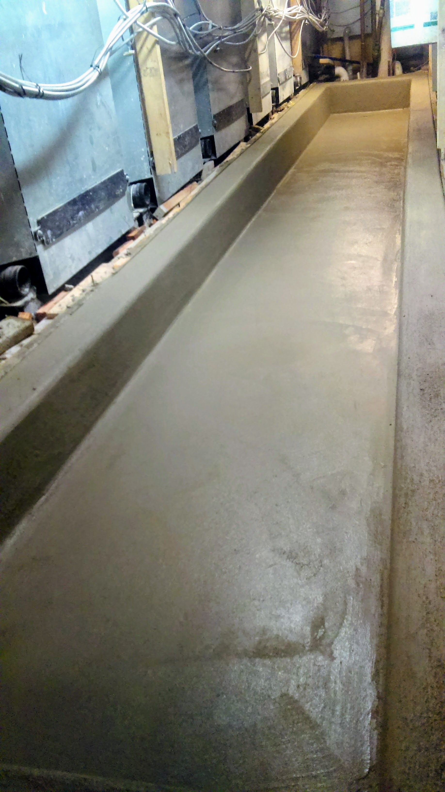 Long, rectangular concrete trench in an industrial setting, next to machinery and a corridor.