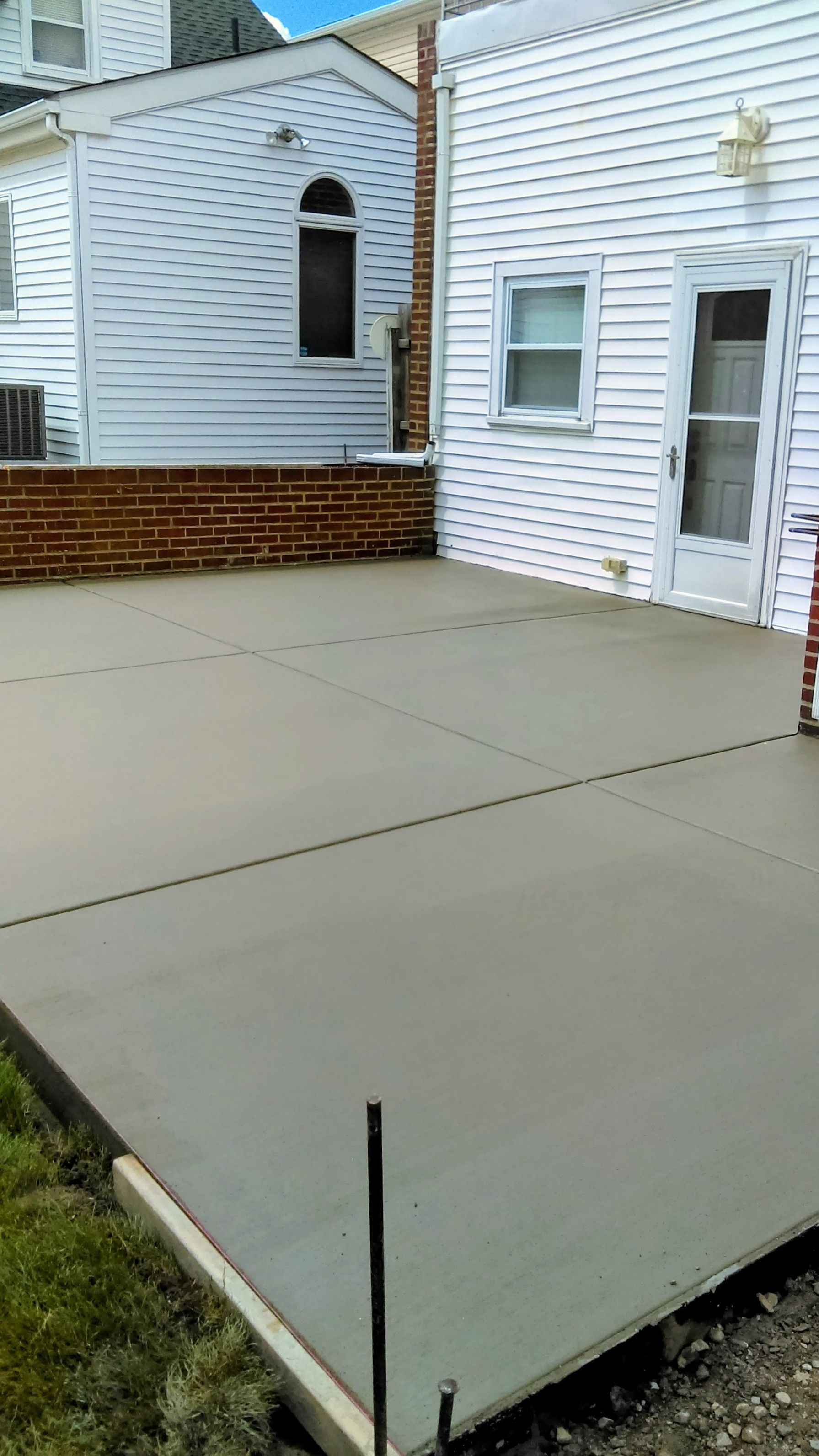 A freshly poured concrete patio next to a brick wall and white house, with green grass in the foreground.